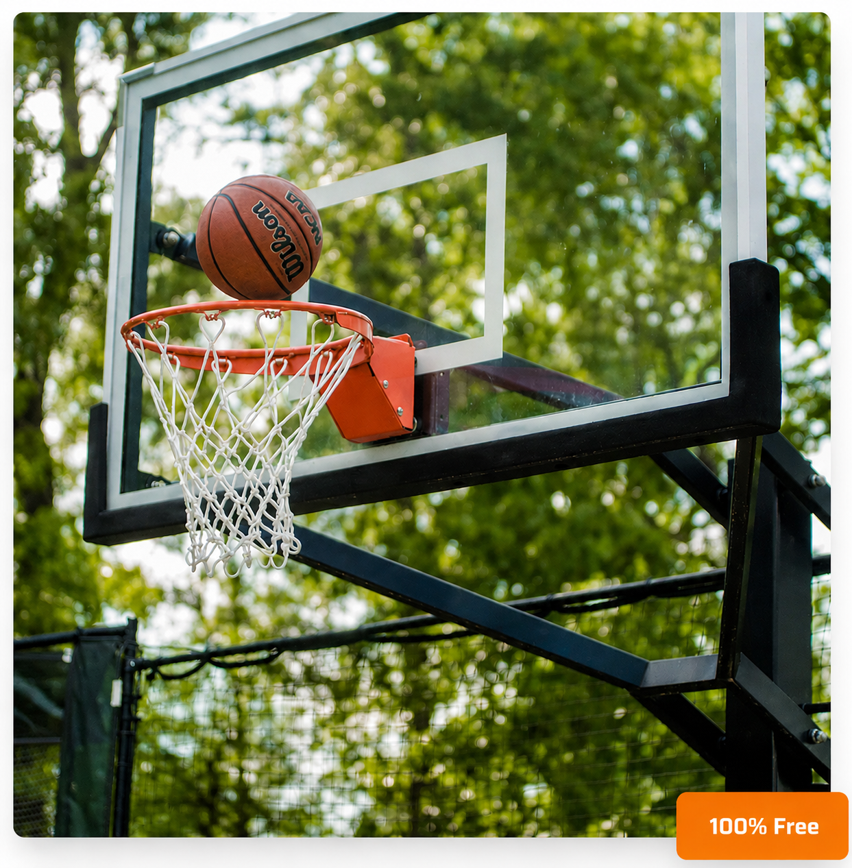A Wilson basketball sits on the rim of an outdoor basketball hoop with a clear backboard and white net against green trees.