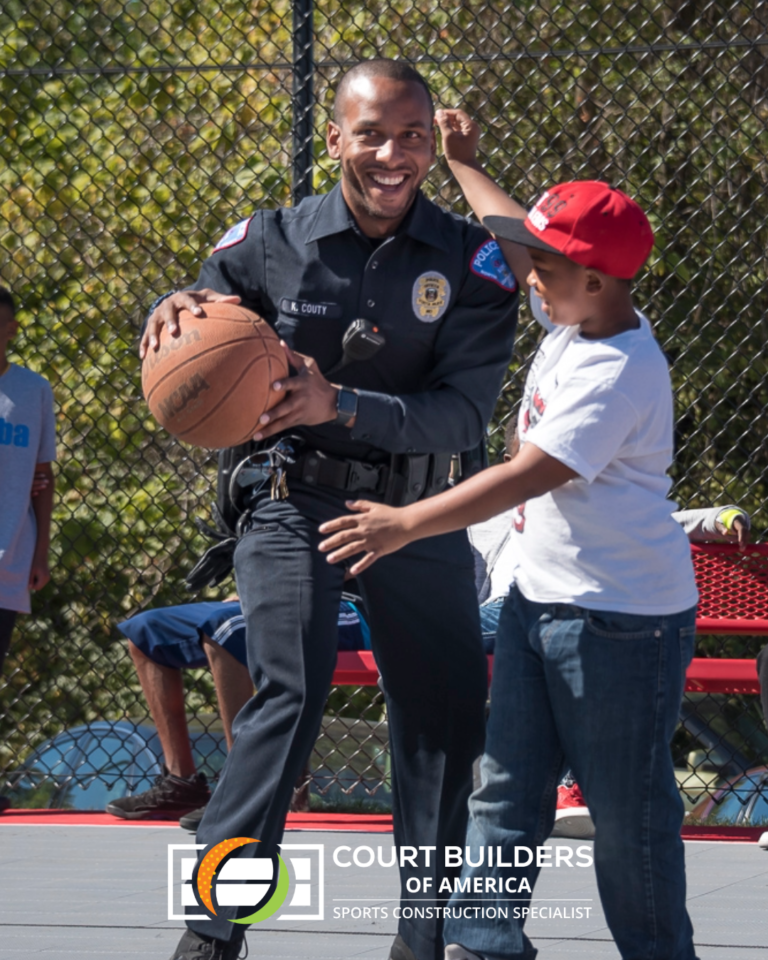 A smiling police officer holding a basketball while interacting with a person on a court in front of a chain-link fence.