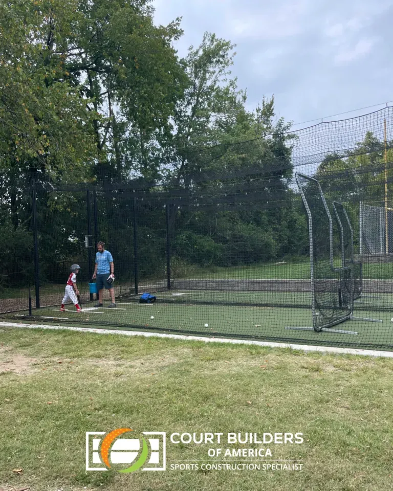 A child practicing baseball with an adult coach in an outdoor netted batting cage on a turf field.