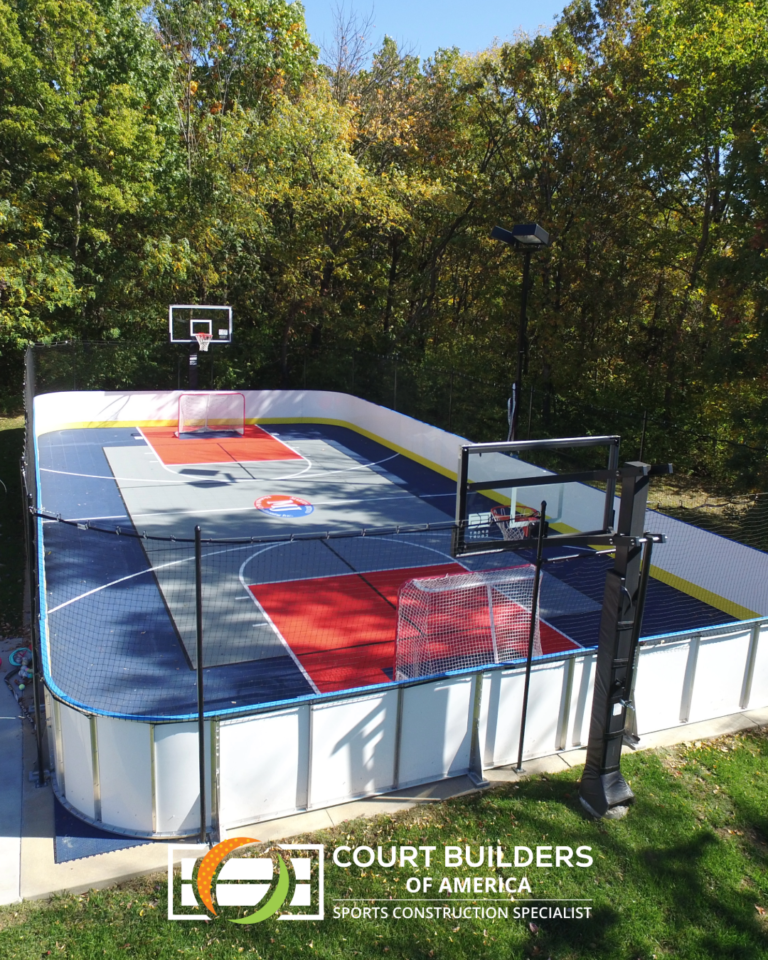 A residential multi-sport court with basketball hoops, hockey goals, and white dasher boards surrounded by trees.