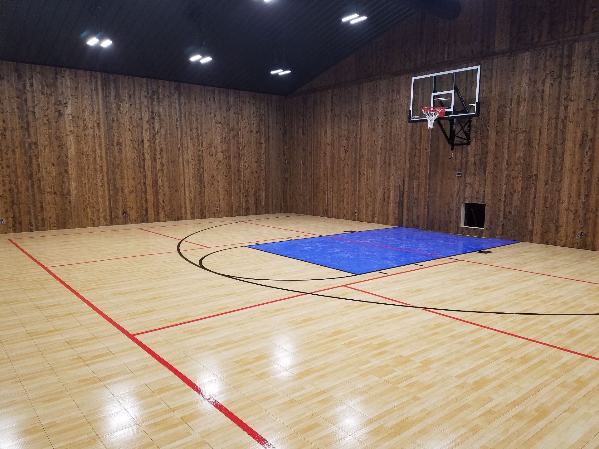 Indoor basketball court with wood-paneled walls, light wood flooring, red court lines, and a blue mat under the hoop.