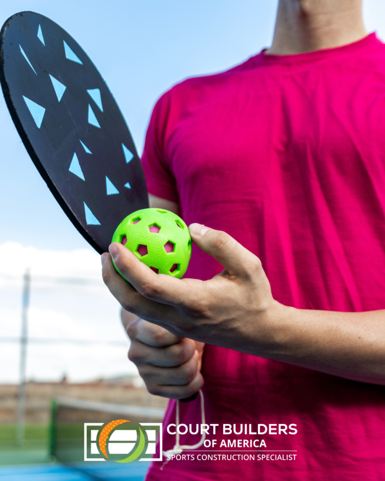 A person in a pink shirt holds a pickleball paddle and a bright green ball on an outdoor court.