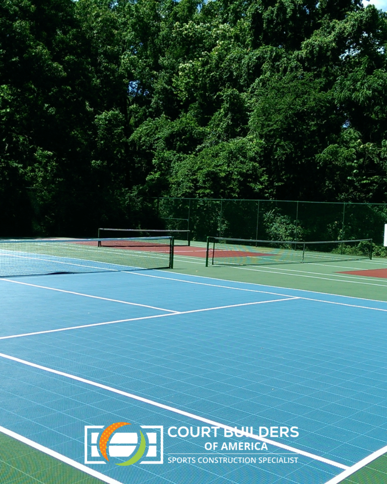 A blue and green modular tennis court set against a backdrop of trees, with a logo overlay for Court Builders of America.