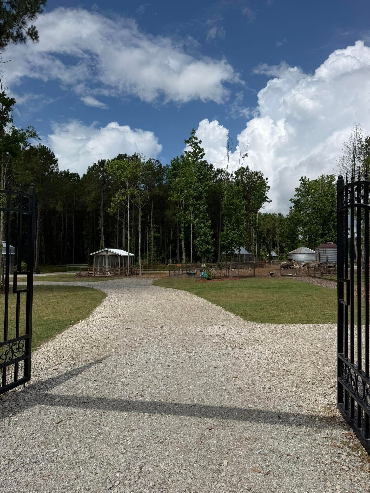 Open wrought-iron gates lead to a gravel path through a grassy clearing and trees under a blue, cloudy sky.