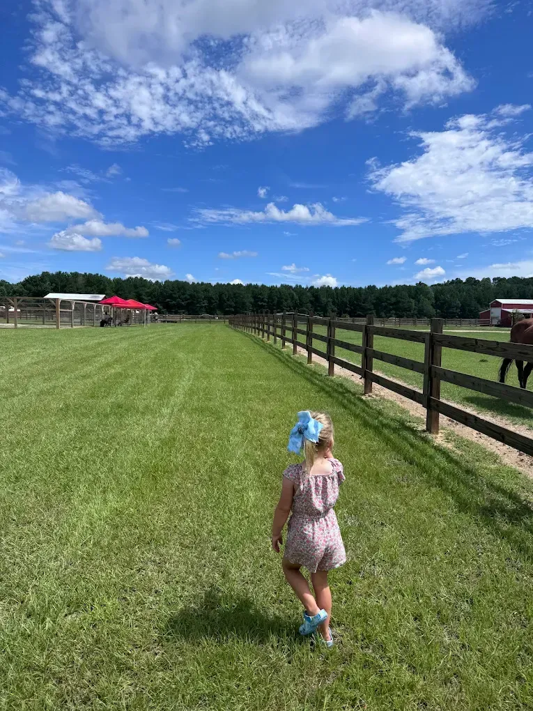 Child running across grassy field beside a fence under a bright blue sky