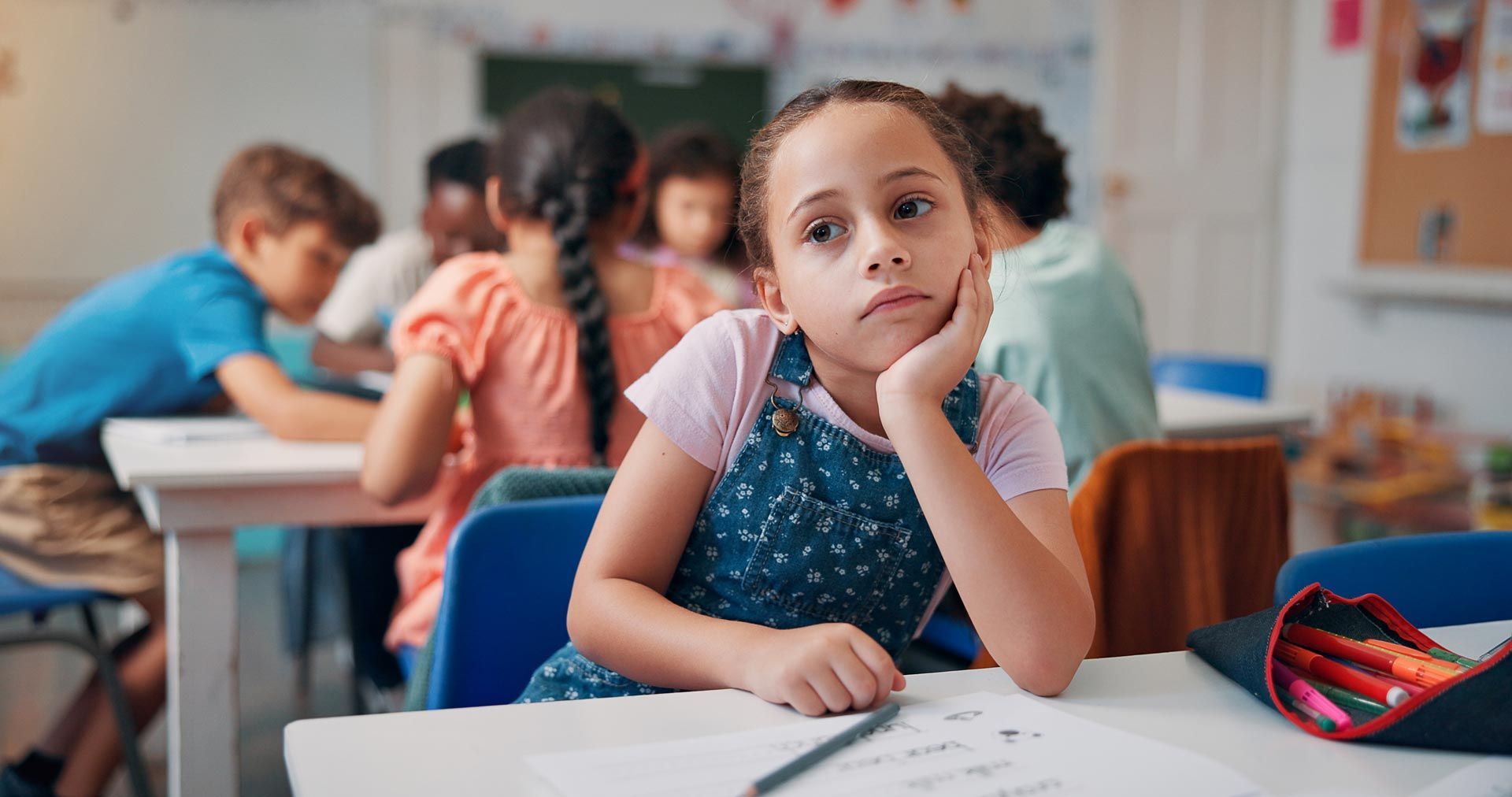 Student sitting at a classroom desk, resting her head on her hand while others work in the background.