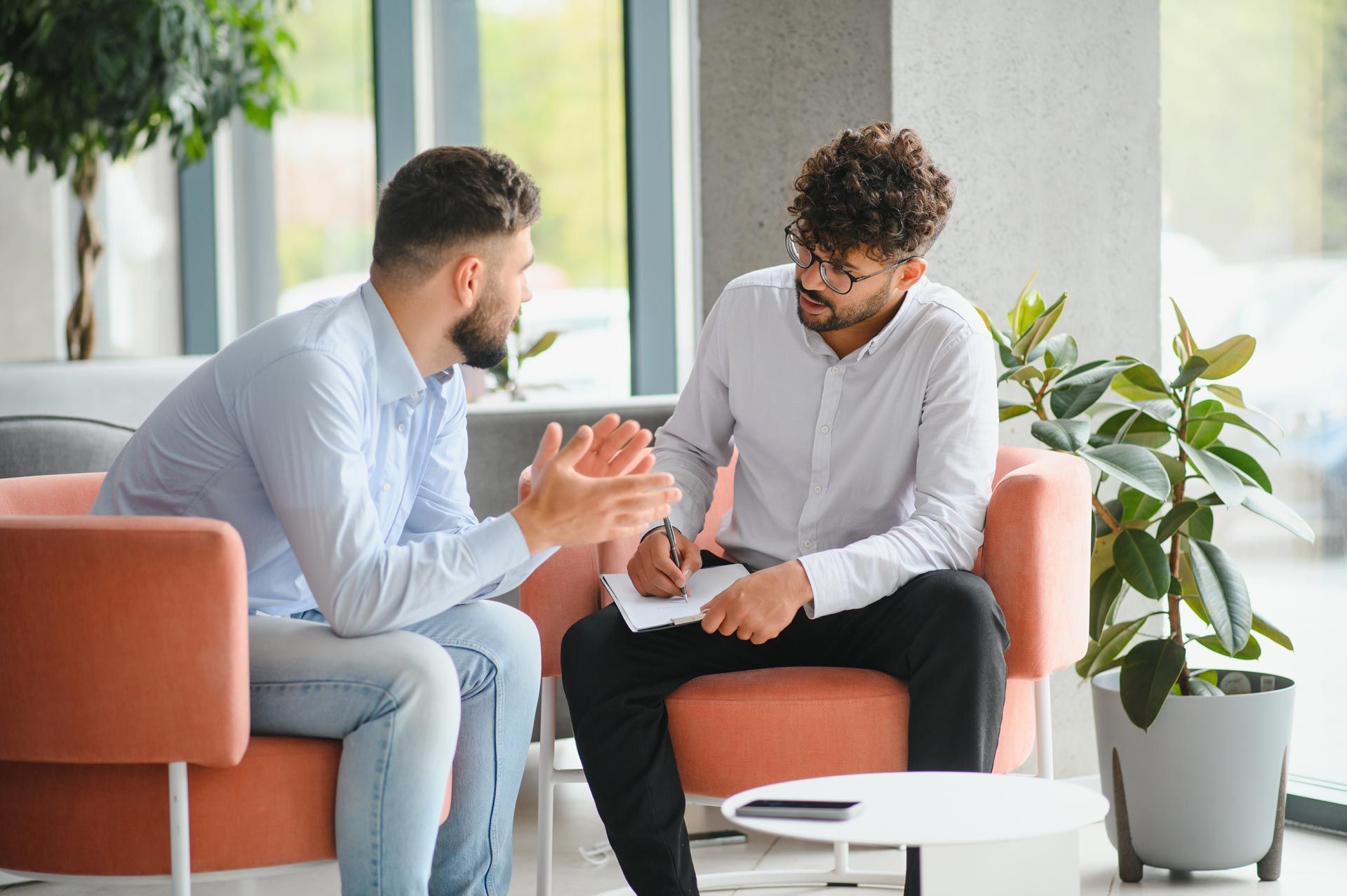 Two men in conversation on an orange sofa in a bright office lounge, one holding a notepad.