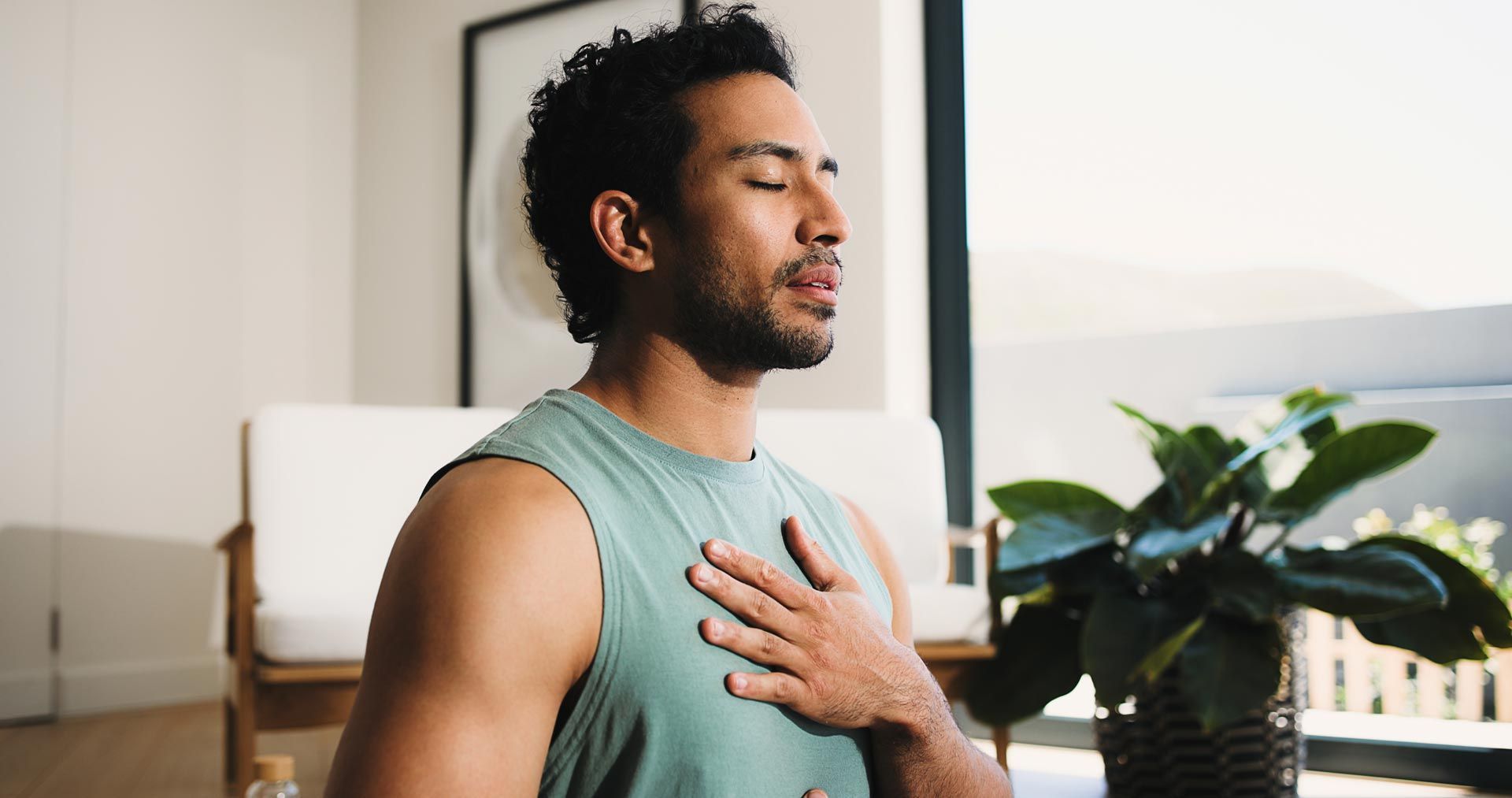 Man in a green tank top holding his chest while breathing calmly indoors near a plant and window.