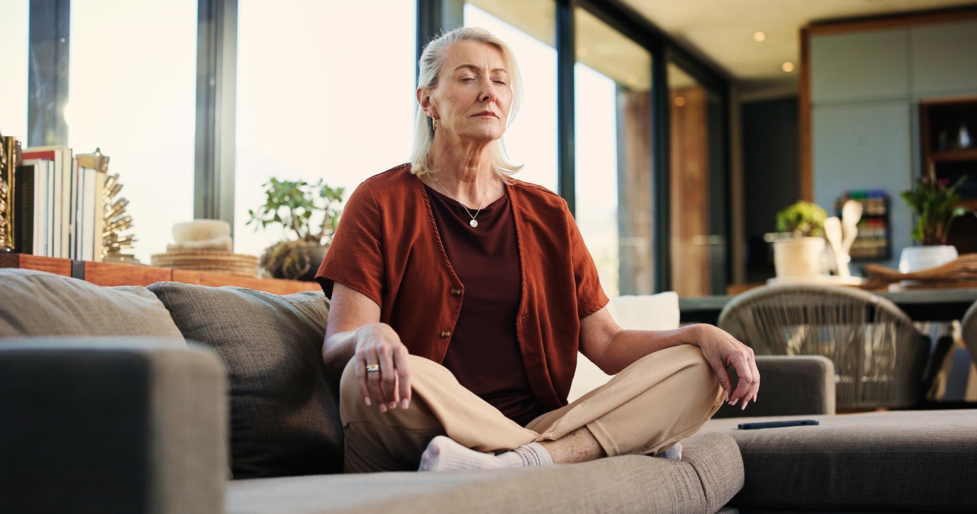 Person sitting cross-legged on a couch in a bright living room, wearing a rust cardigan and beige pants.