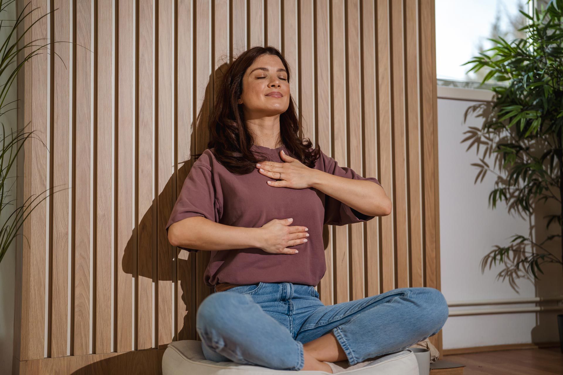 Woman meditating cross-legged on a cushion in a wooden room, hand on chest and abdomen.