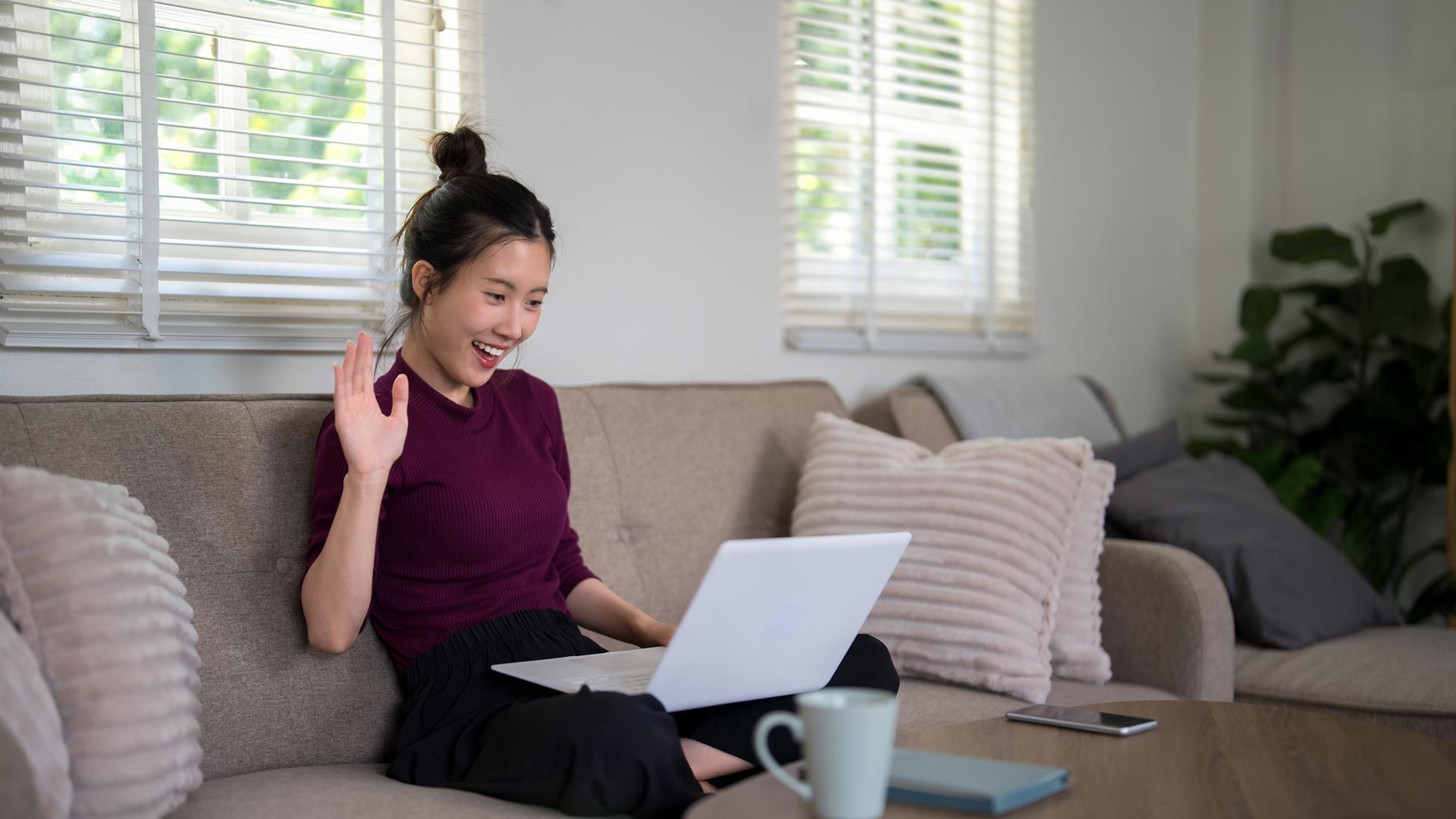 Person video chatting on a laptop in a bright living room, smiling and waving from a couch