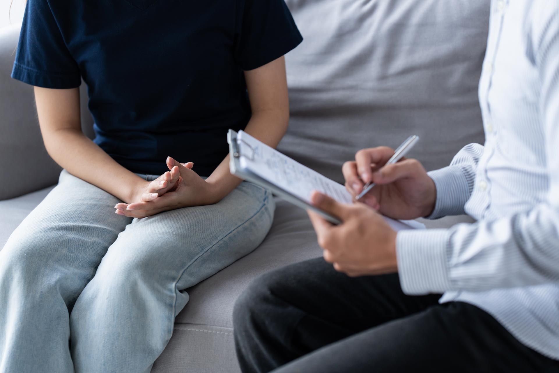Two people sit on a couch during a conversation, one taking notes on a clipboard.