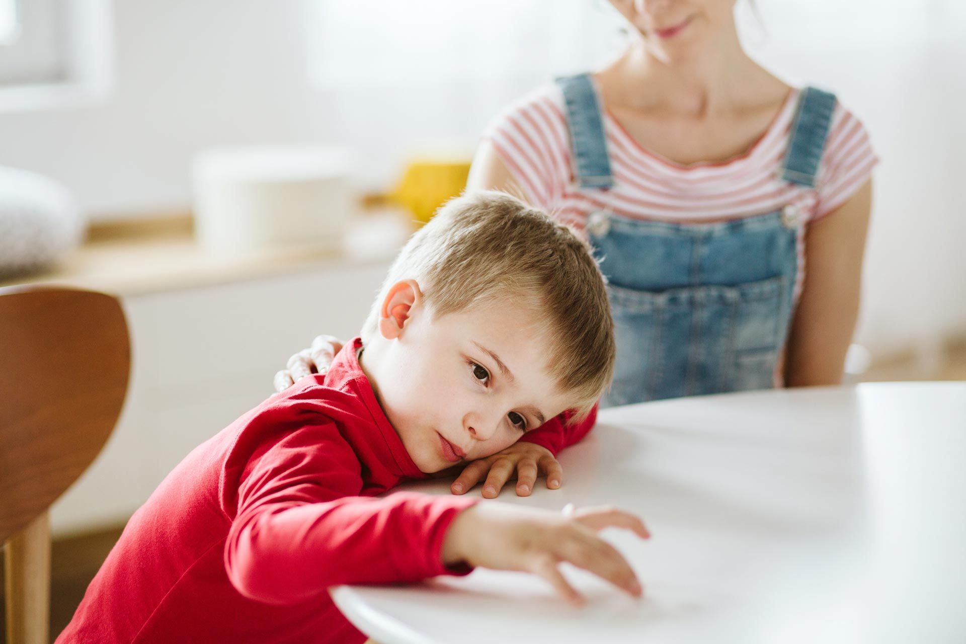Child in a red shirt resting on a table, with an adult in overalls behind them in a bright room