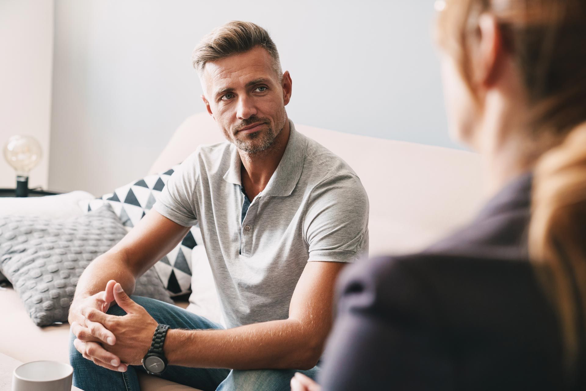 Two people talking on a couch in a bright living room, one listening attentively.