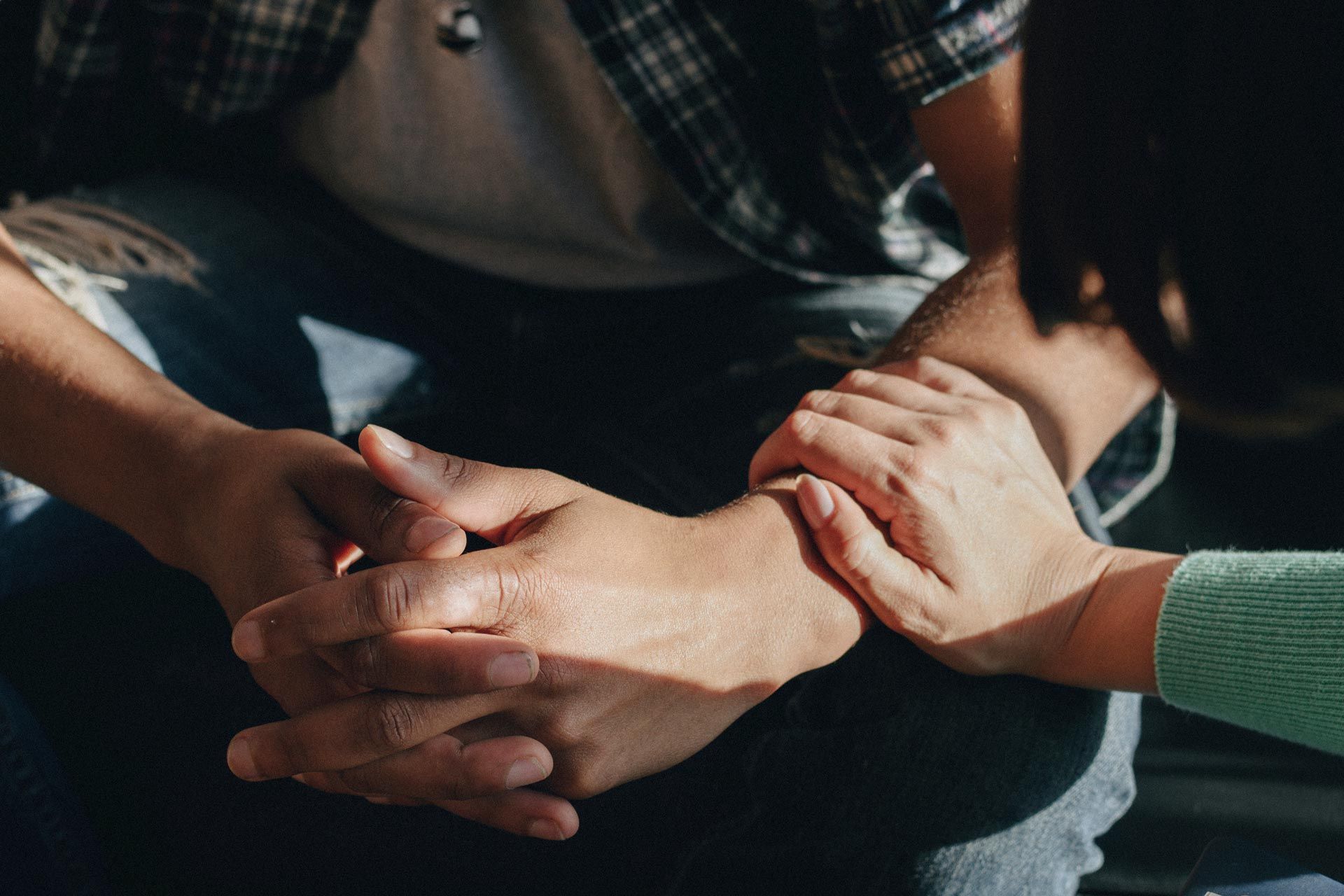 Two hands clasped together in a supportive gesture on a seated person's lap, with another hand resting on top.