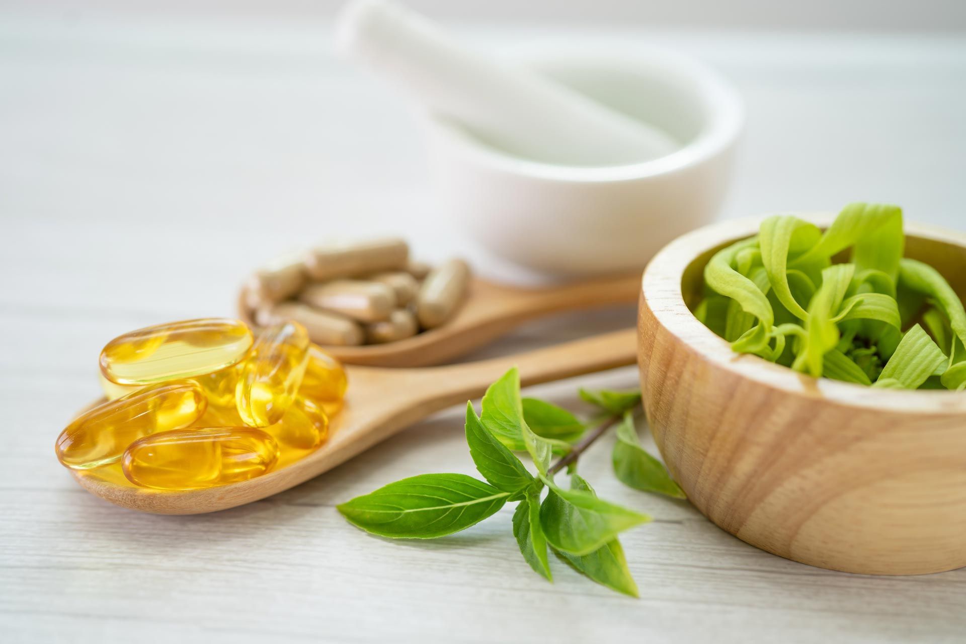 Wooden spoon with yellow capsules, herbs, and fresh basil beside a bowl of greens on a white table