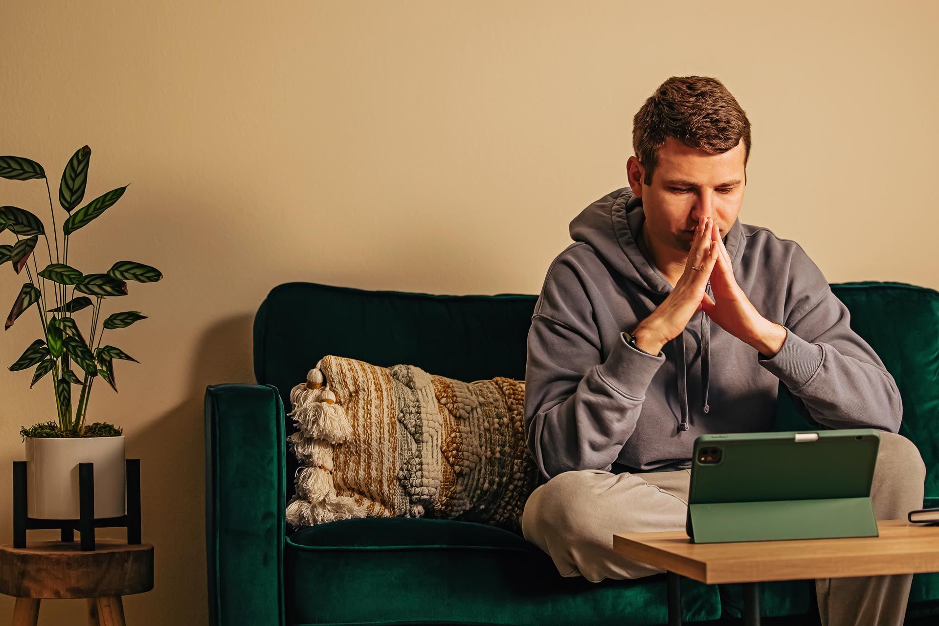 Man sitting on a green couch, hands clasped in thought, looking at a laptop in a cozy living room.