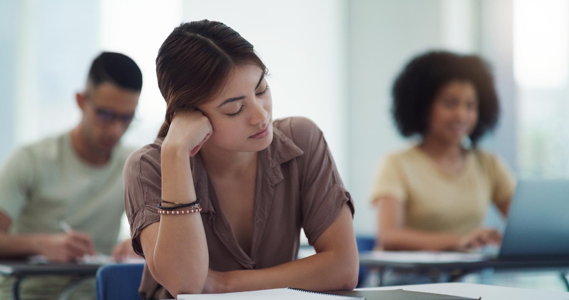 Student leaning on hand at a classroom desk, looking down while others work in the background.