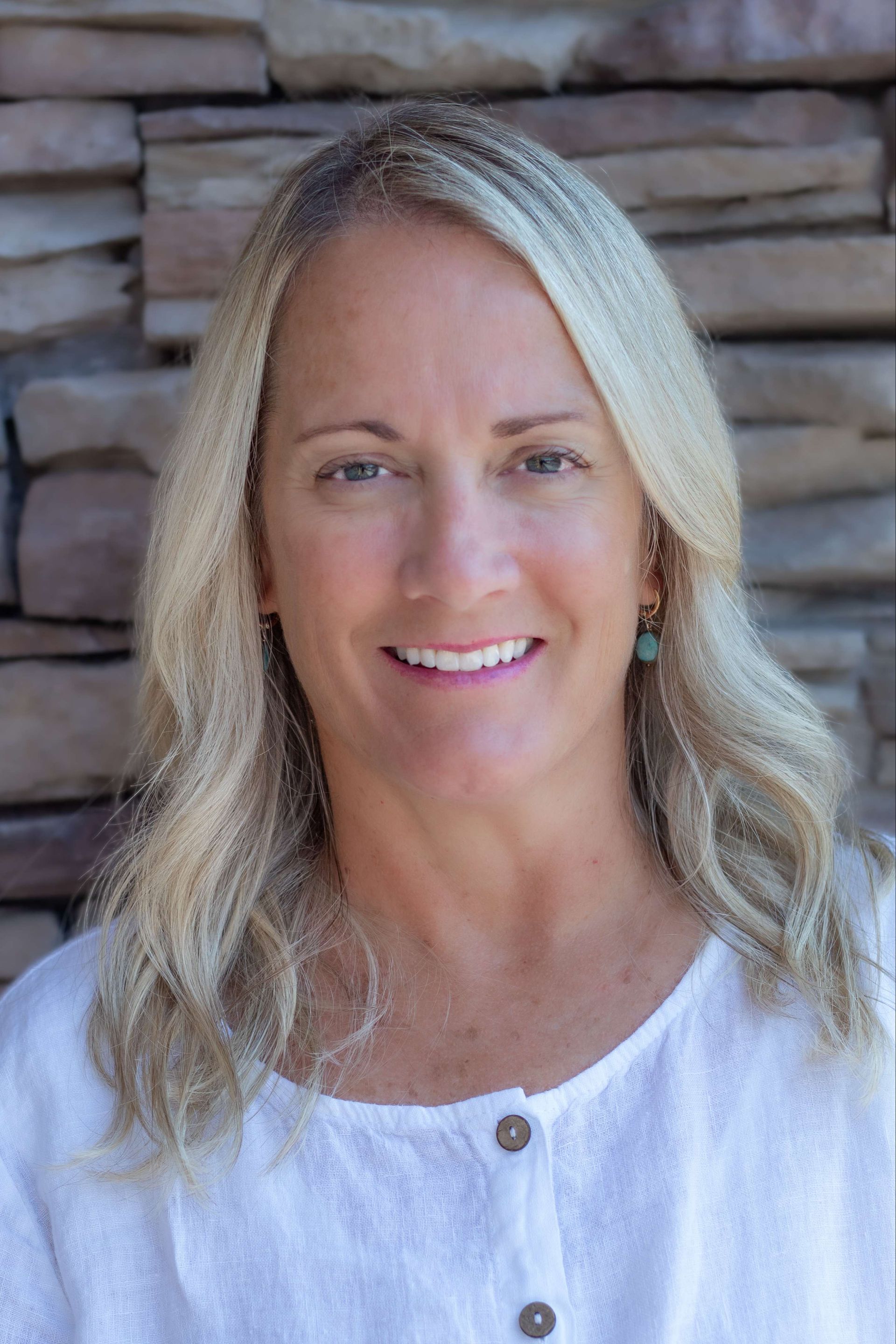 Smiling blonde woman in a white top standing against a stone wall