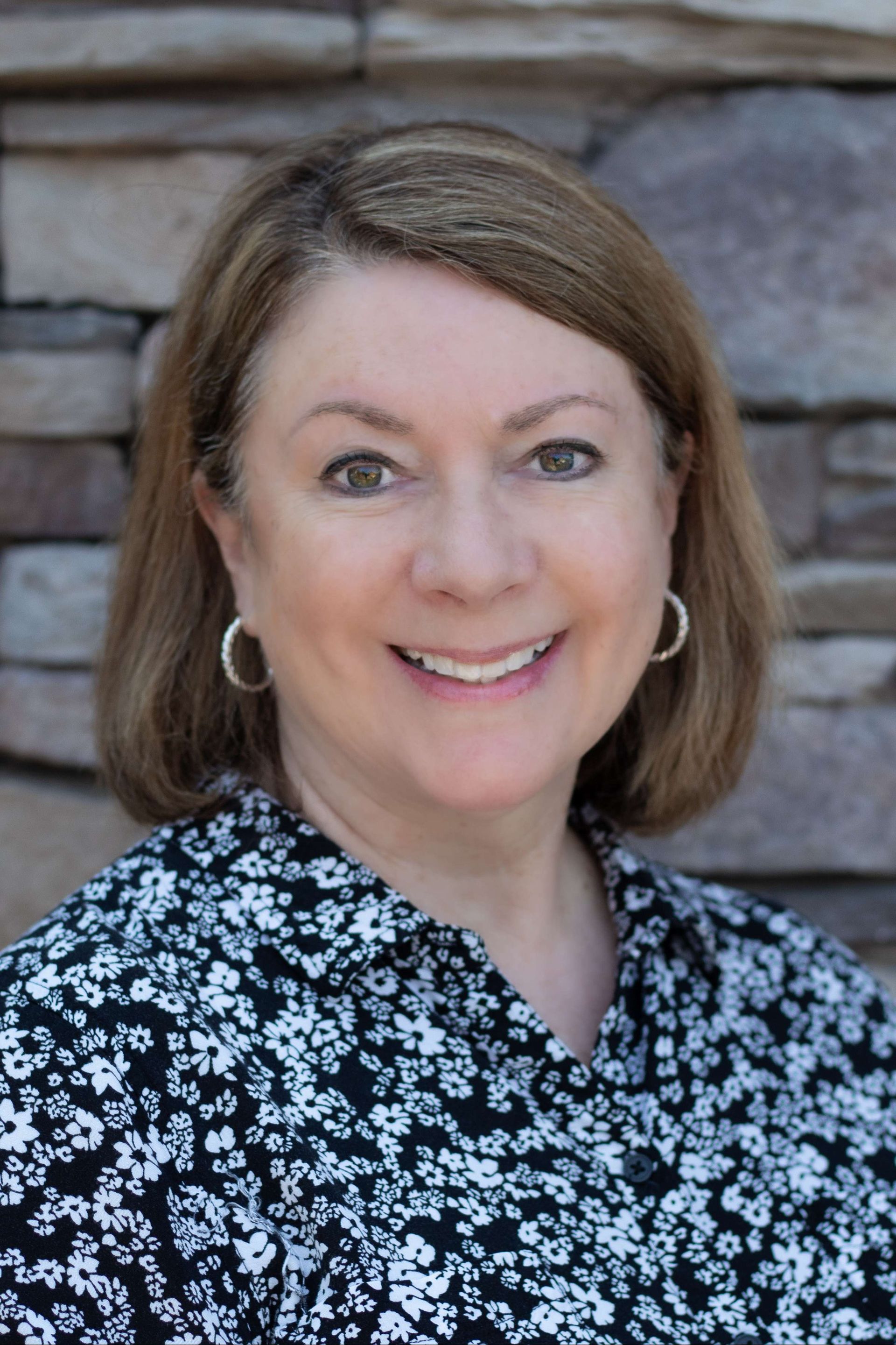 Smiling person in a black floral blouse posed against a stone wall.
