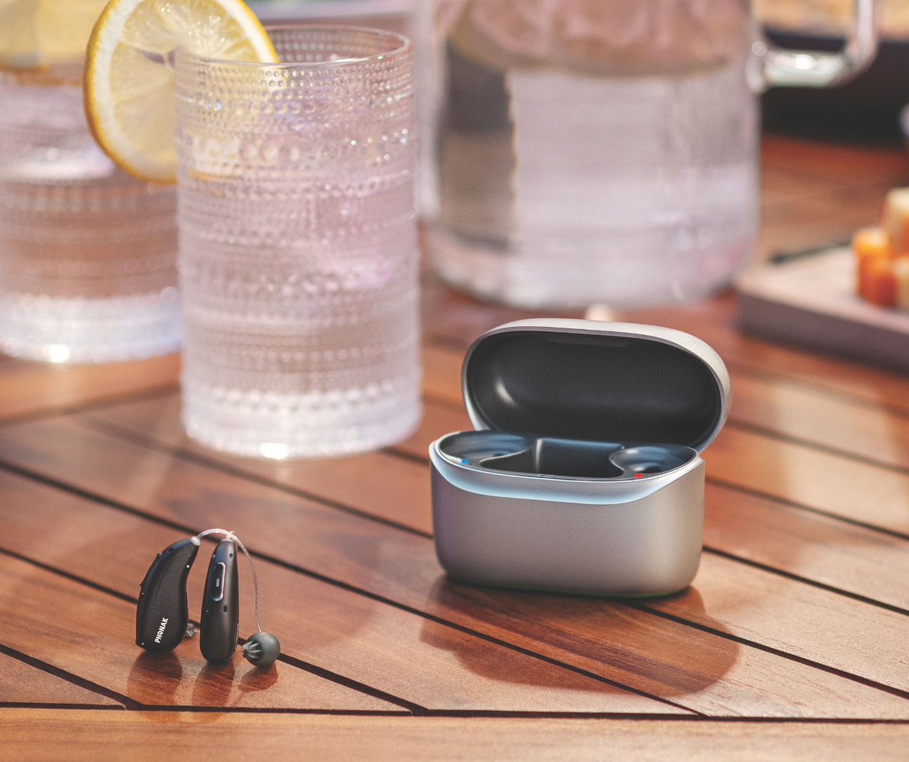 Wireless earbuds in an open charging case on a wooden table beside glasses and a lemon drink