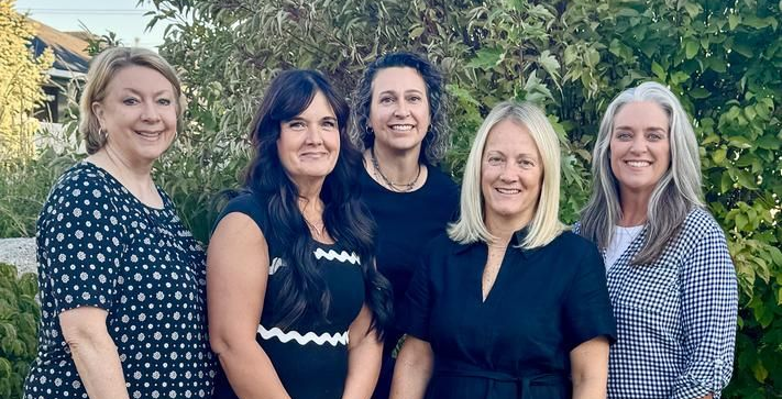 Five women standing outdoors, smiling in front of greenery and a stone wall.