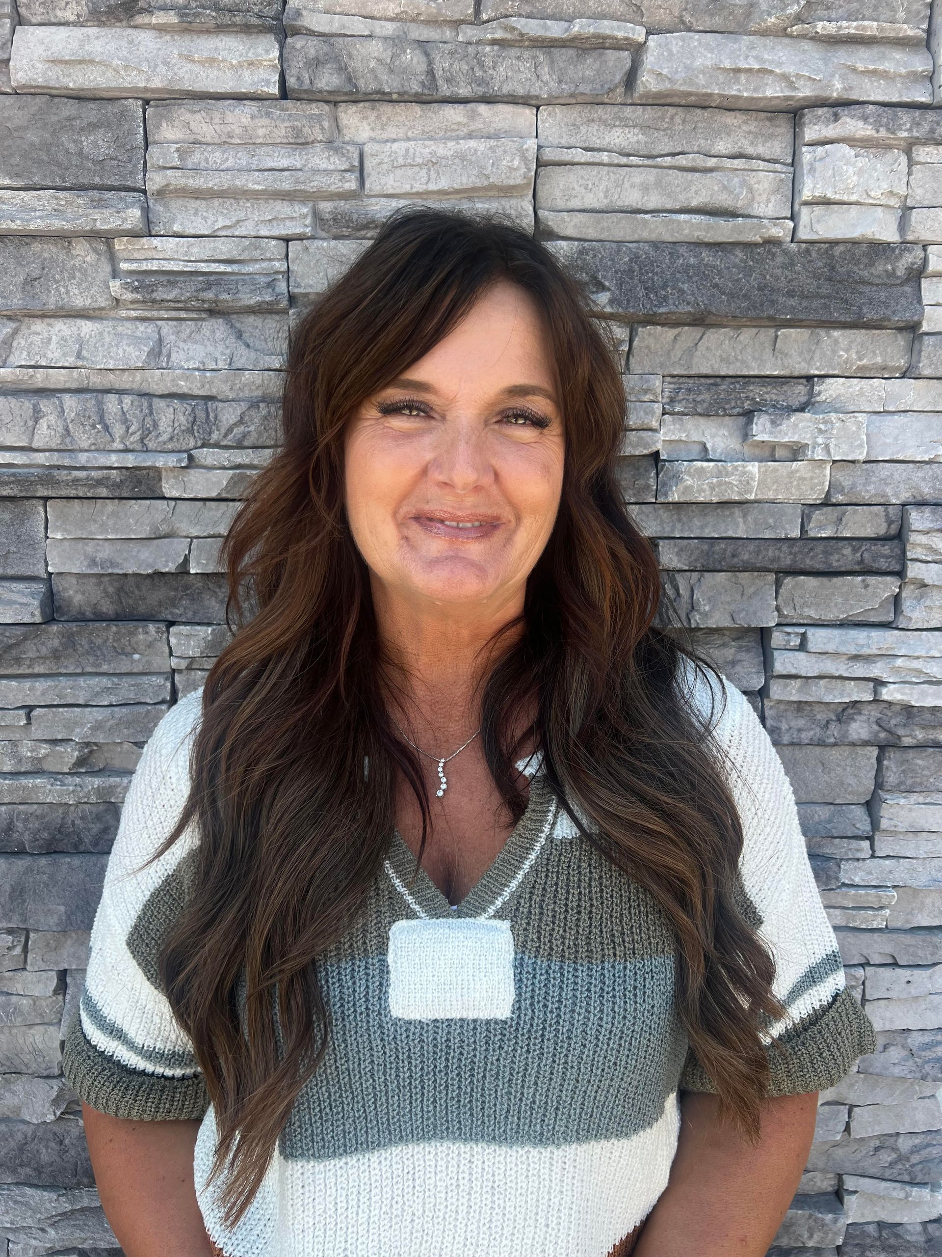 Woman with long dark hair standing against a stacked stone wall, wearing a striped sweater and necklace.