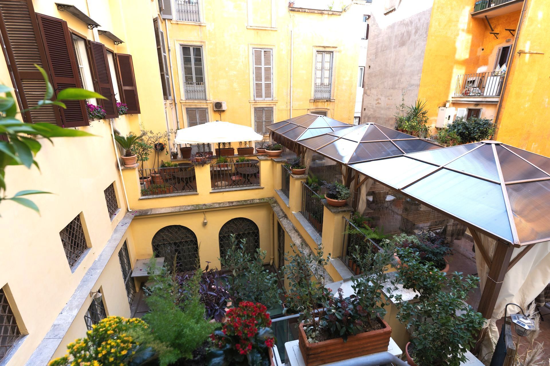 Innenhof mit gelben Gebäuden, Pflanzen, überdachten Sitzgelegenheiten und einem weißen Sonnenschirm.