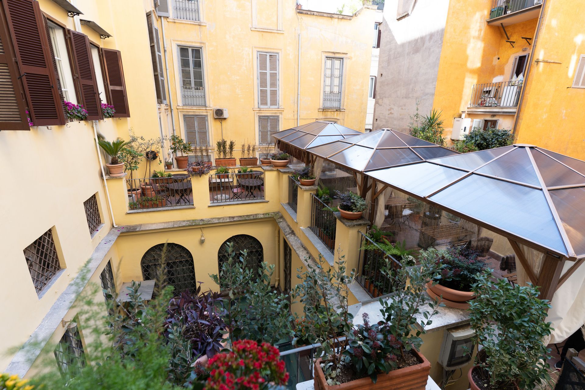Innenhof mit gelben Gebäuden, Pflanzen, Topfblumen und überdachten Sitzgelegenheiten.