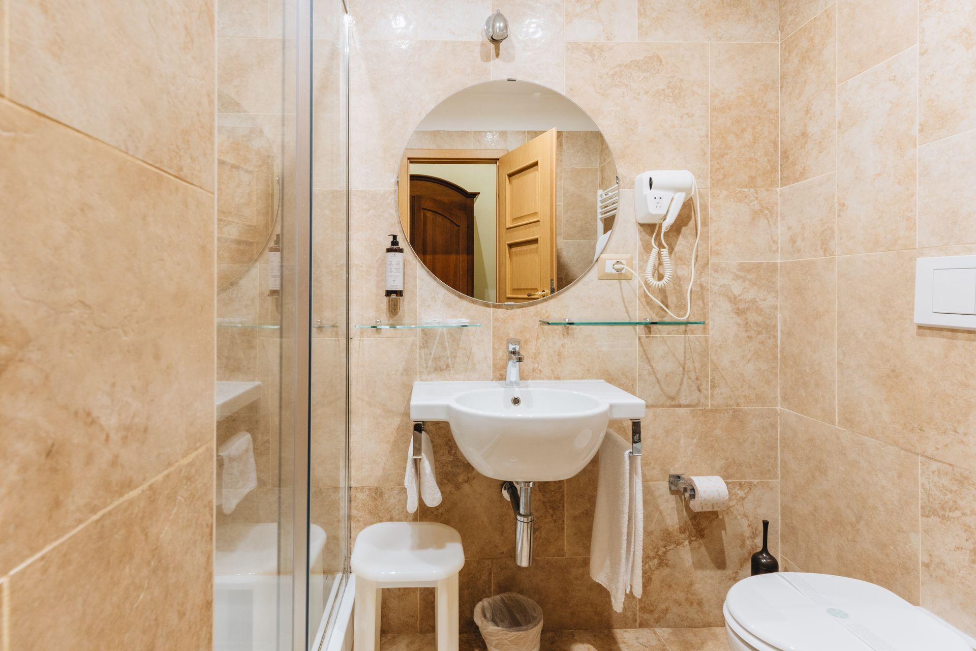 Kleines Badezimmer mit rundem Spiegel, Waschbecken, Toilette und Dusche. Beige geflieste Wände.
