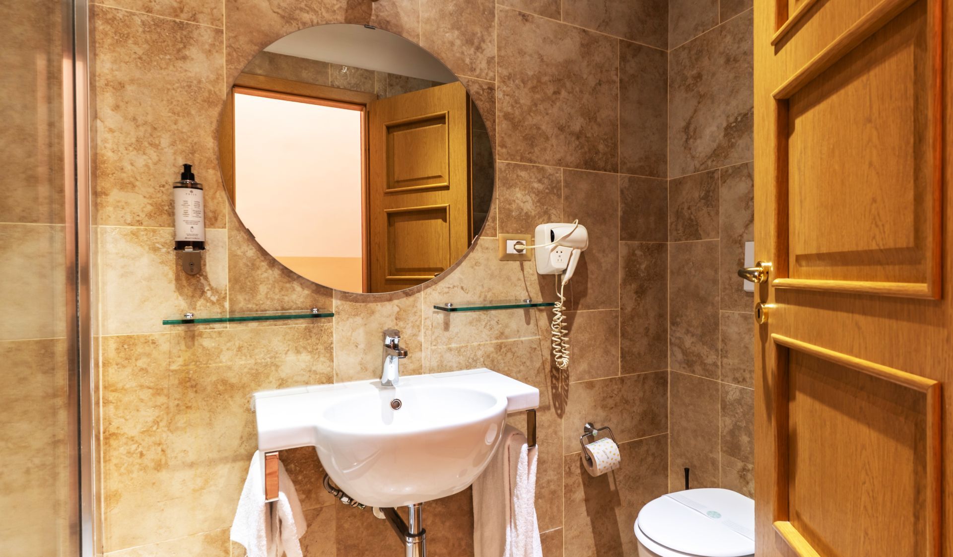 Badezimmer mit rundem Spiegel, Waschbecken, Toilette und hell gefliesten Wänden.