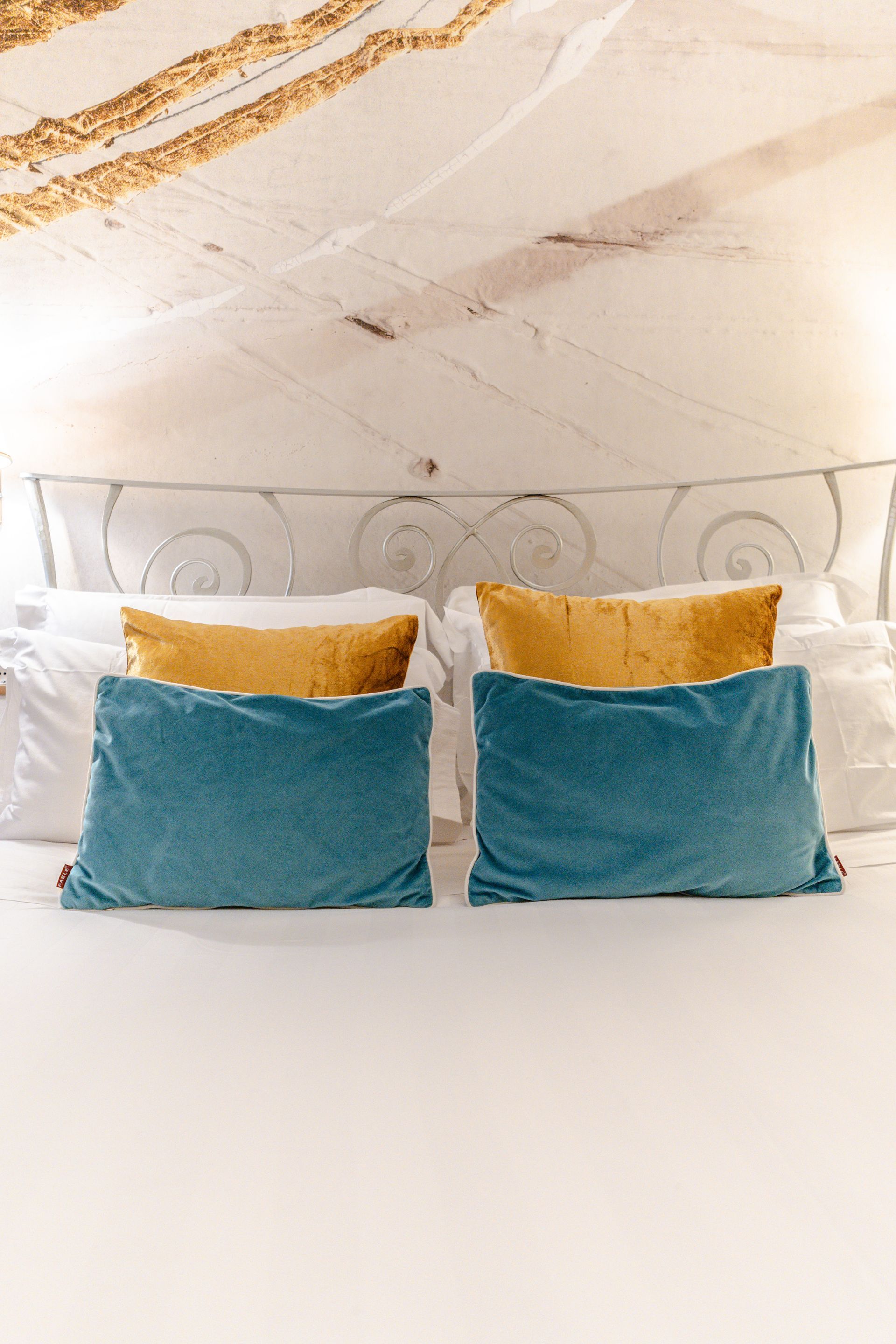 Bett mit weißen Laken, blau-goldenen Samtkissen und verziertem weißen Kopfteil.