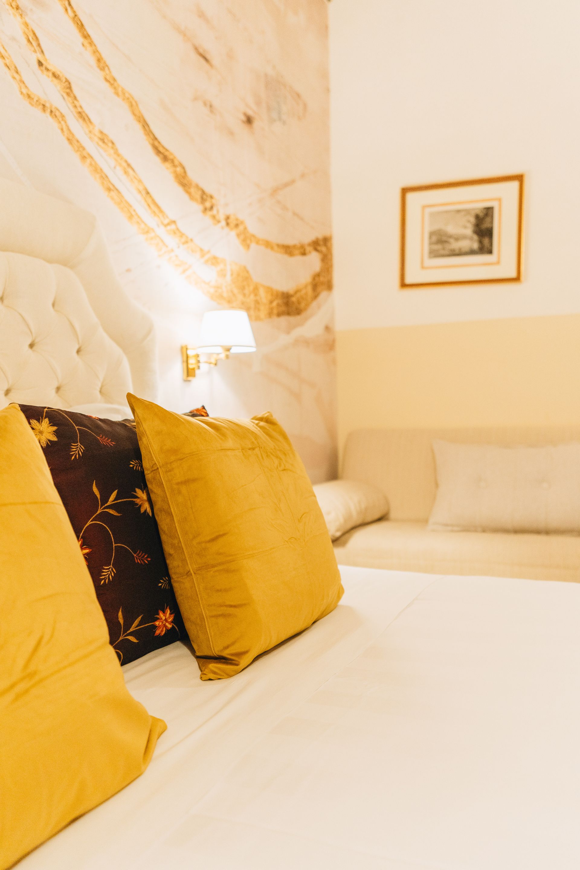 Gemütliches Schlafzimmer mit weißem Bett, goldenen Kissen und gemusterter Wand, über dem Tagesbett hängt ein kleines Gemälde.