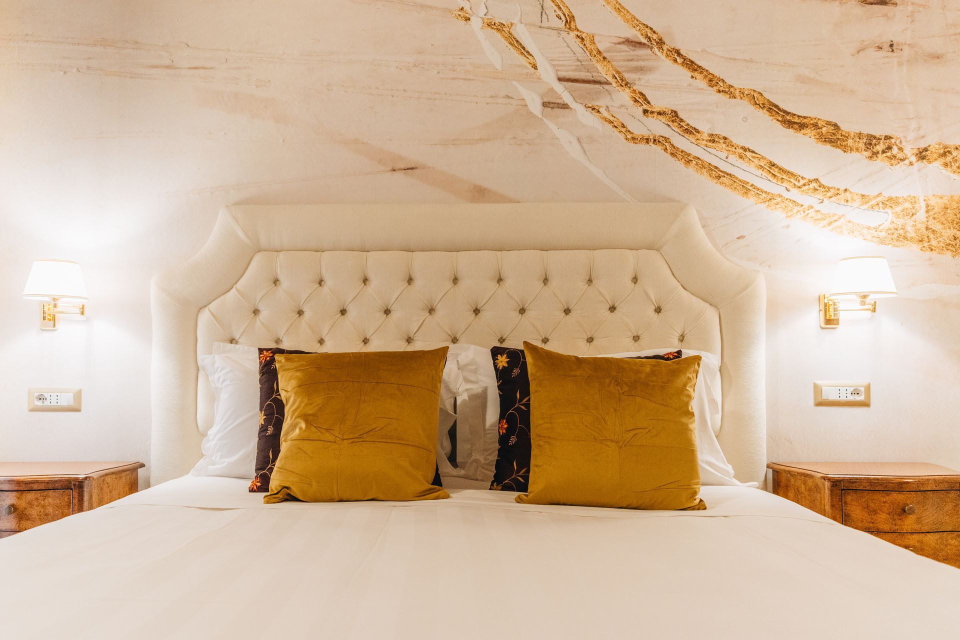 Schlafzimmer mit weißem, gestepptem Kopfteil, goldenen Kissen und Nachttischen, sanfte Beleuchtung.