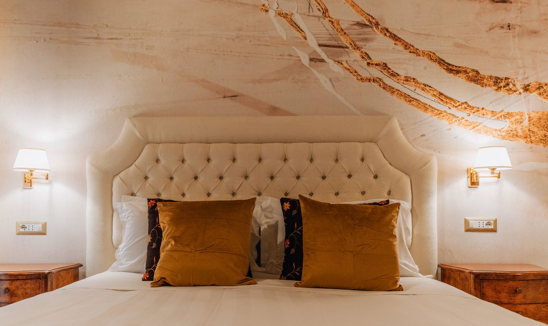 Schlafzimmer mit gestepptem Kopfteil, goldfarbenen Kissen, Nachttischen, Wandleuchten und abstrakter Deckenmalerei.