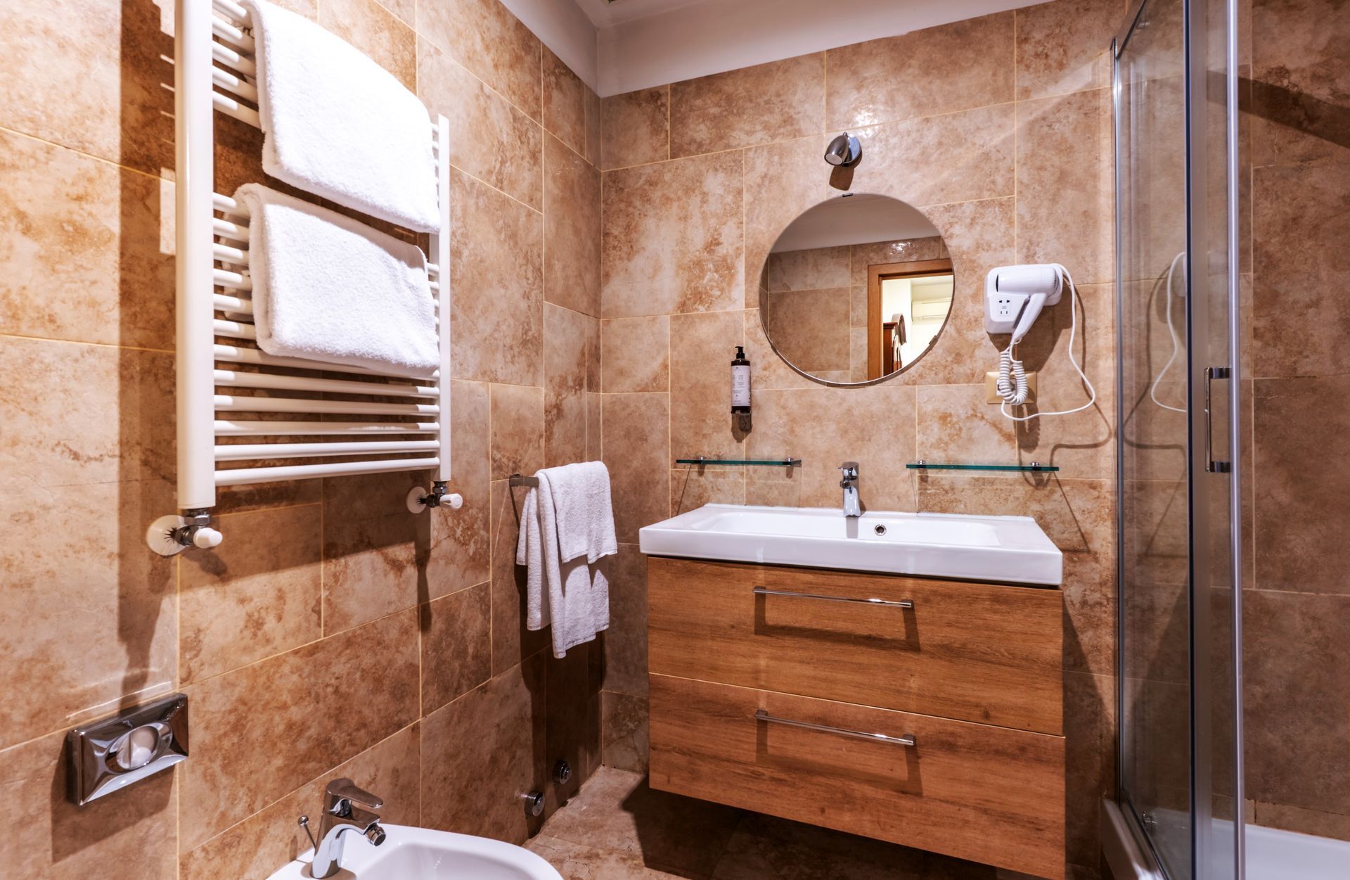 Badezimmer mit gefliesten Wänden, Waschtisch aus Holz, rundem Spiegel, Dusche und Handtuchwärmer.