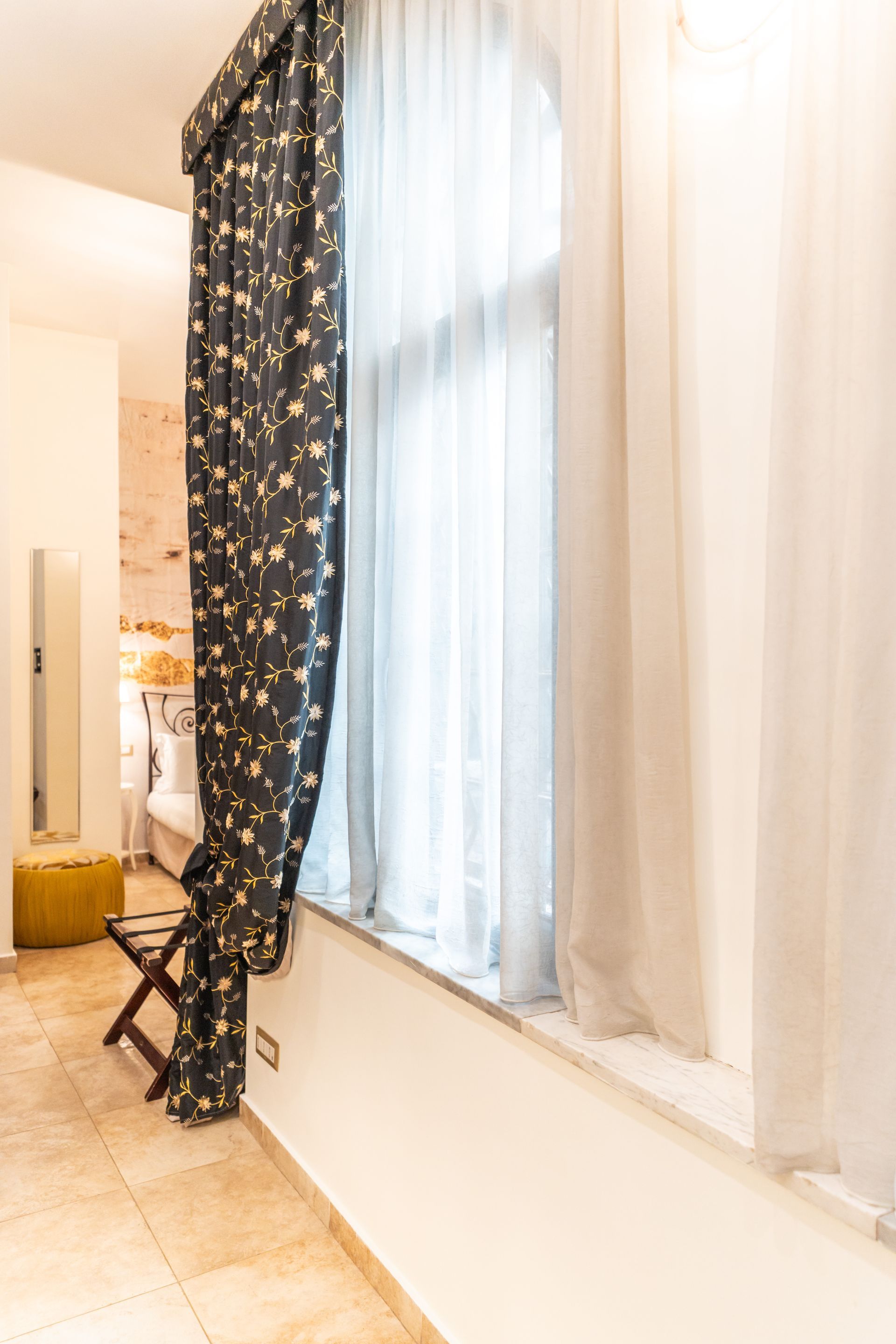 Ein dunkler, geblümter Vorhang und ein durchsichtiger weißer Vorhang rahmen ein hohes Fenster mit tiefer Fensterbank ein.
