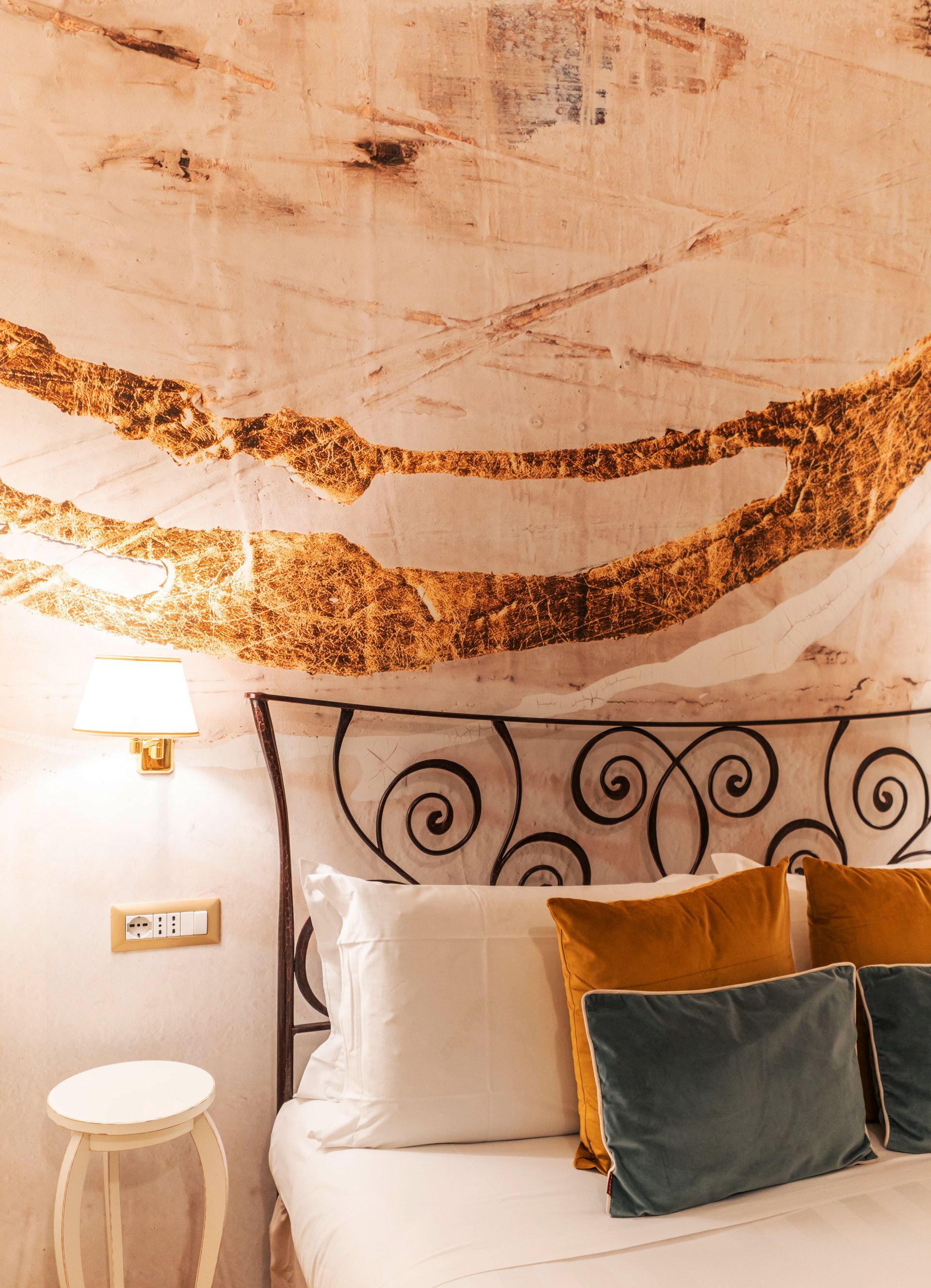 Schlafzimmer mit dekorativer Wandgestaltung in Gold und Beige, schmiedeeisernem Kopfteil, Kissen und kleinem Tisch.