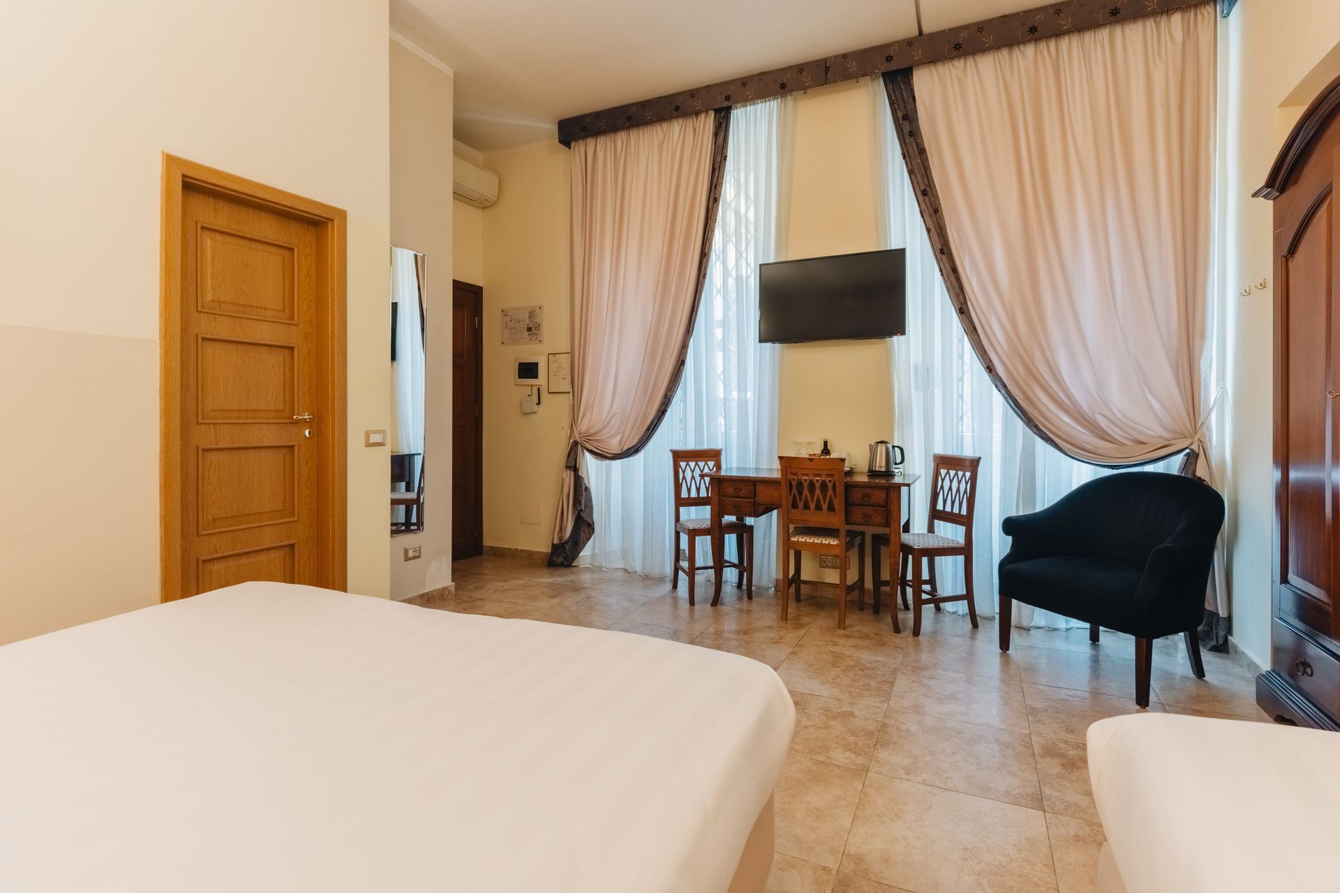 Hotelzimmer-Interieur mit Bett, Tisch, Stühlen, Fernseher und großen Fenstern mit Vorhängen.