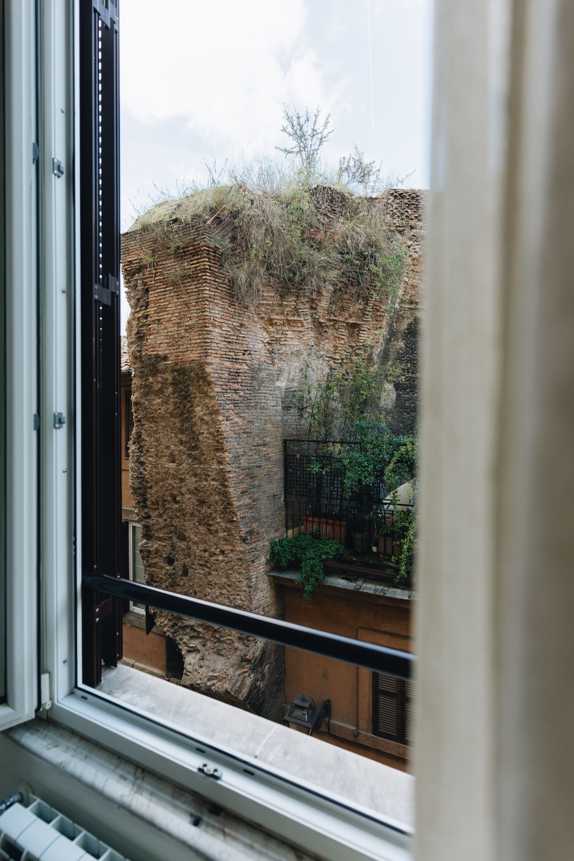 Blick aus dem Fenster: verwittertes Backsteingebäude mit Vegetation, eingerahmt von Fenster und Vorhang.
