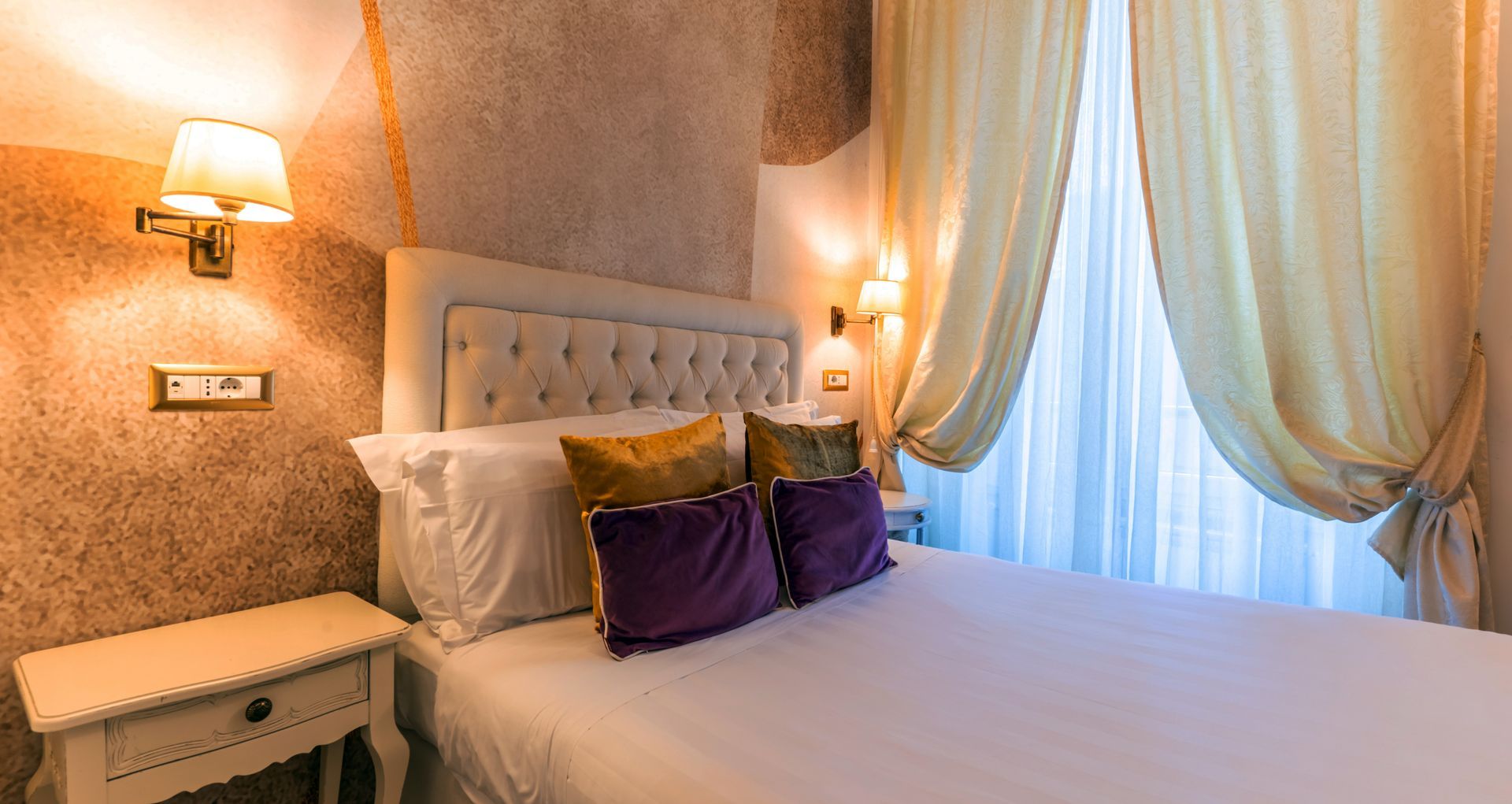 Schlafzimmer mit gestepptem Kopfteil, weißer Bettwäsche, goldenen und violetten Kissen und kunstvollen Wandleuchten.