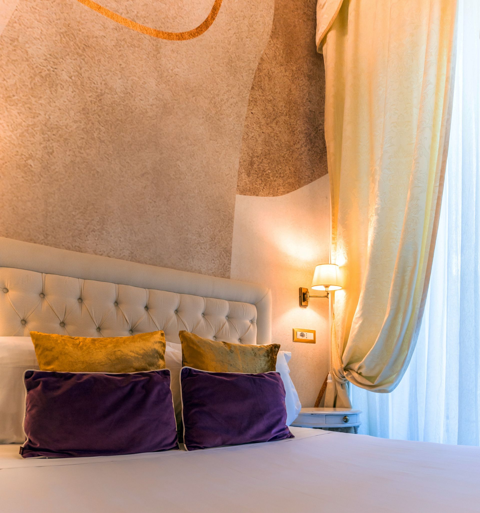 Schlafzimmer mit weißem, gestepptem Kopfteil, weißer Bettwäsche und lila-goldenen Kissen. Hellbeige Wände.