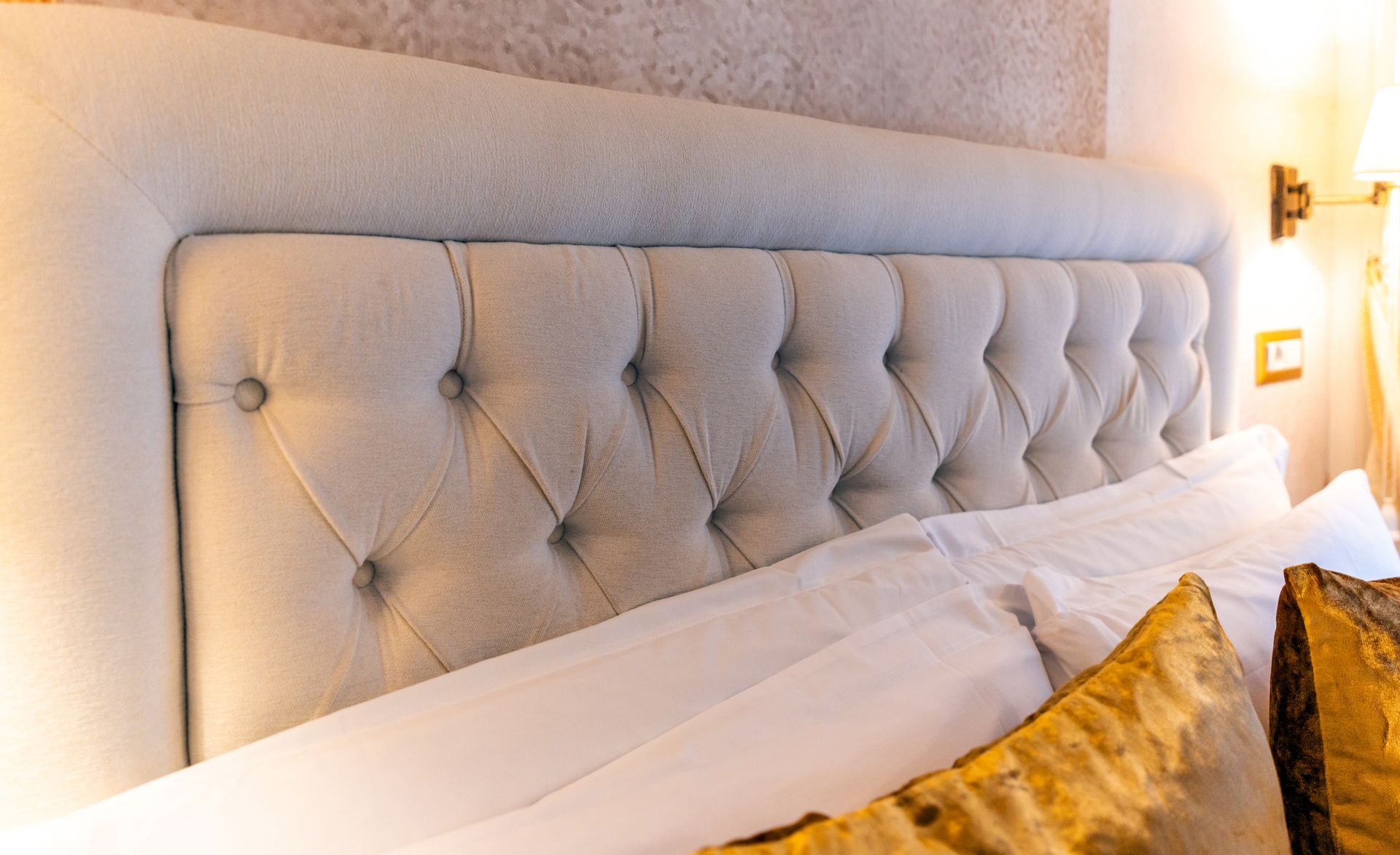 Gepolstertes Kopfteil über einem Bett mit weißen Laken und goldenen Kissen.
