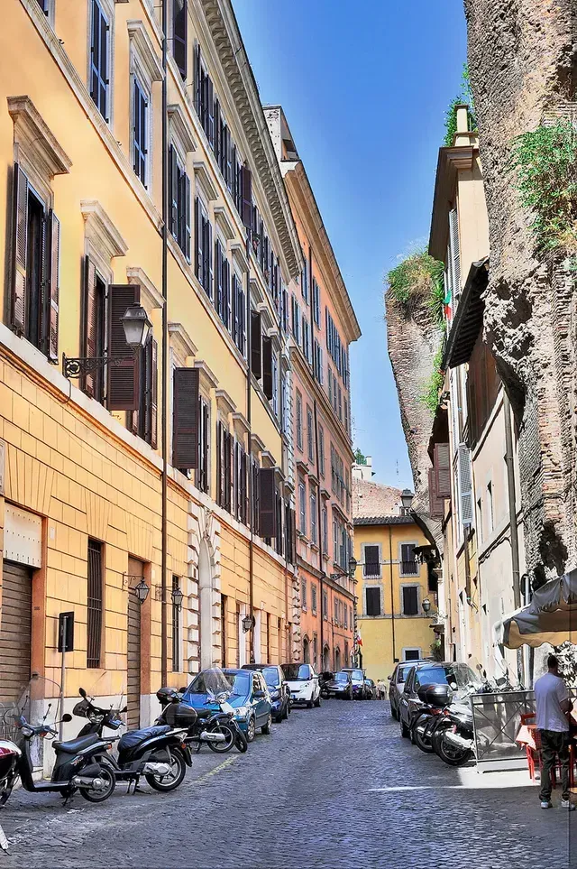 Kopfsteinpflasterstraße in Rom, Italien, gesäumt von Gebäuden, geparkten Autos und Motorrädern. Blauer Himmel darüber.