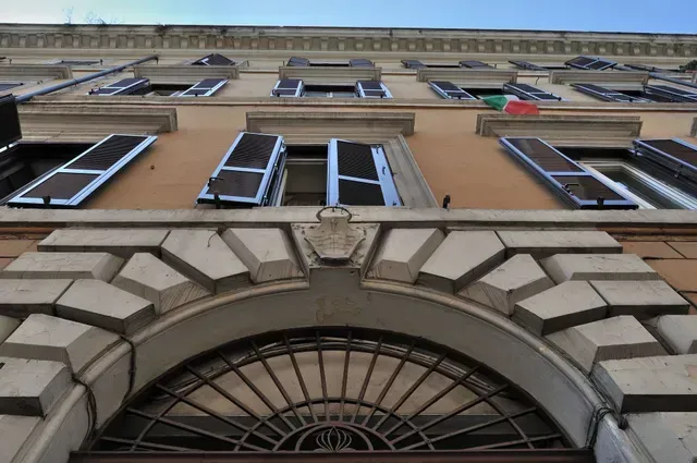 Ansicht von unten auf ein beigefarbenes Gebäude mit Bogeneingang und blauen Fensterläden unter klarem Himmel.