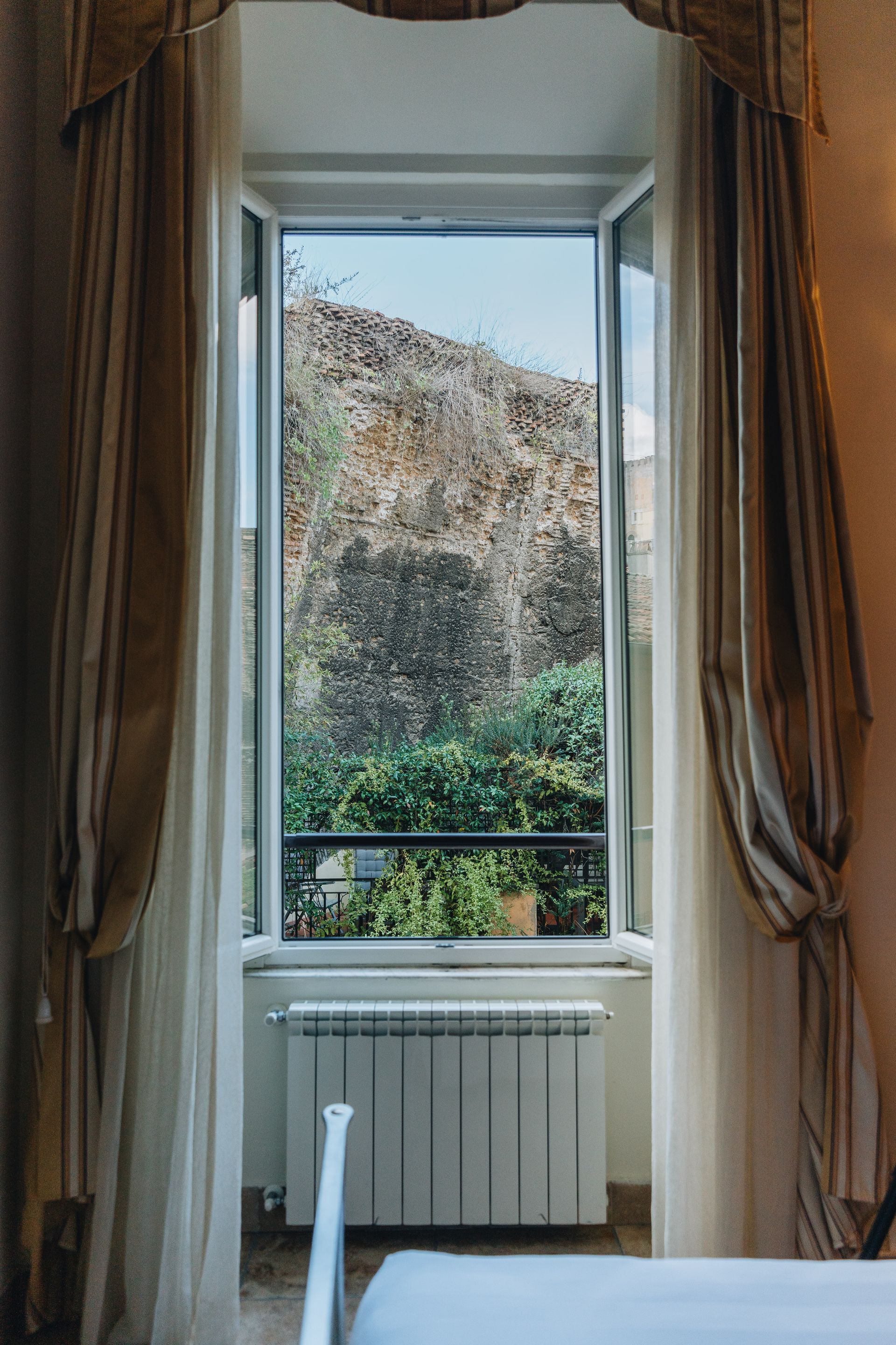 Offenes Fenster mit durchsichtigen Vorhängen, Blick auf einen felsigen Hügel und Grünanlagen, darunter ein Heizkörper.