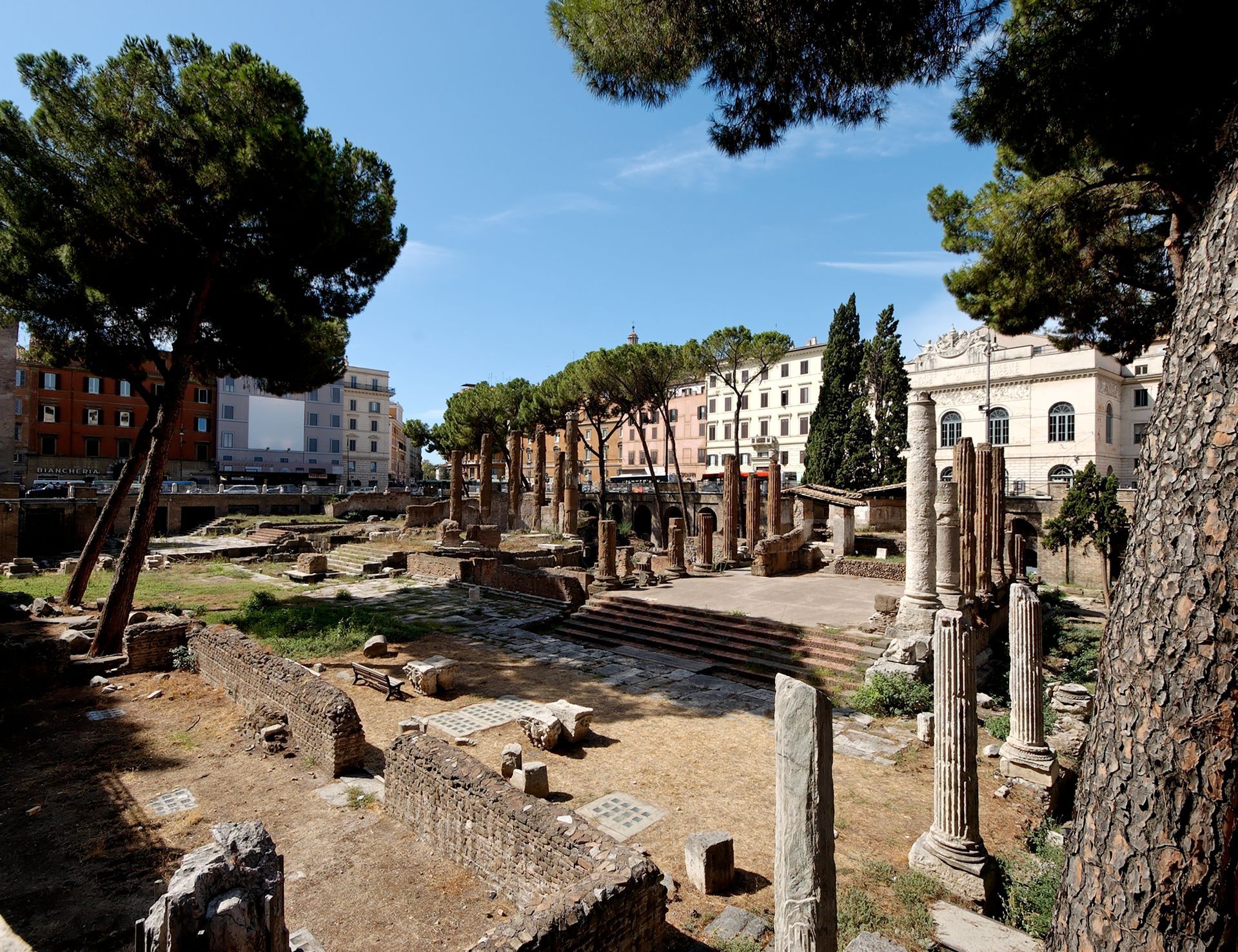 Ruines d'un ancien temple romain dans un cadre urbain ensoleillé, avec colonnes, arbres et bâtiments.