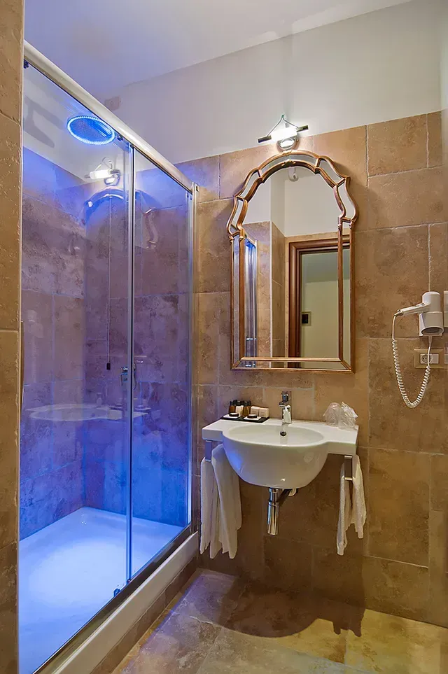Badezimmer mit Glasdusche, Waschbecken, Spiegel und beigefarbenen Fliesen an Wänden und Boden.