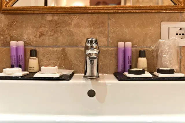 Waschbecken mit Armatur, Toilettenartikeln und Spiegel. Beigefarbene Fliesen im Hintergrund.