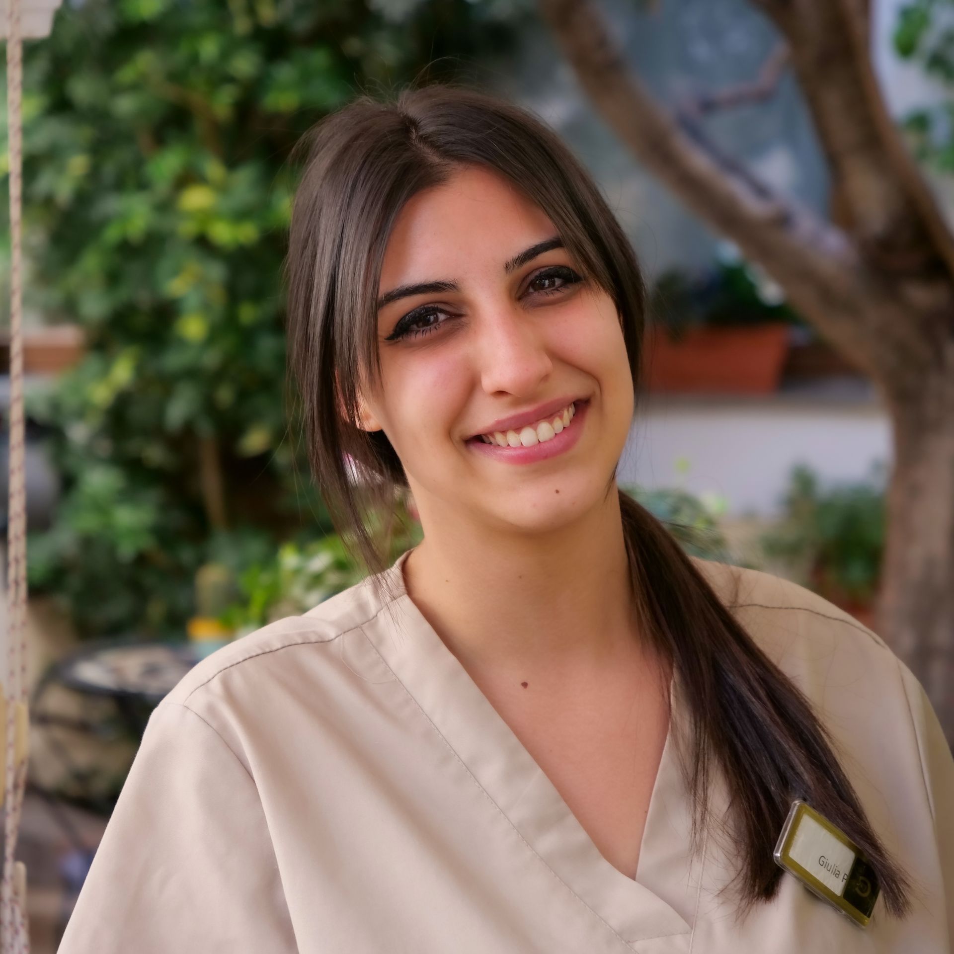 Woman with a ponytail, smiling in a tan uniform, near greenery.